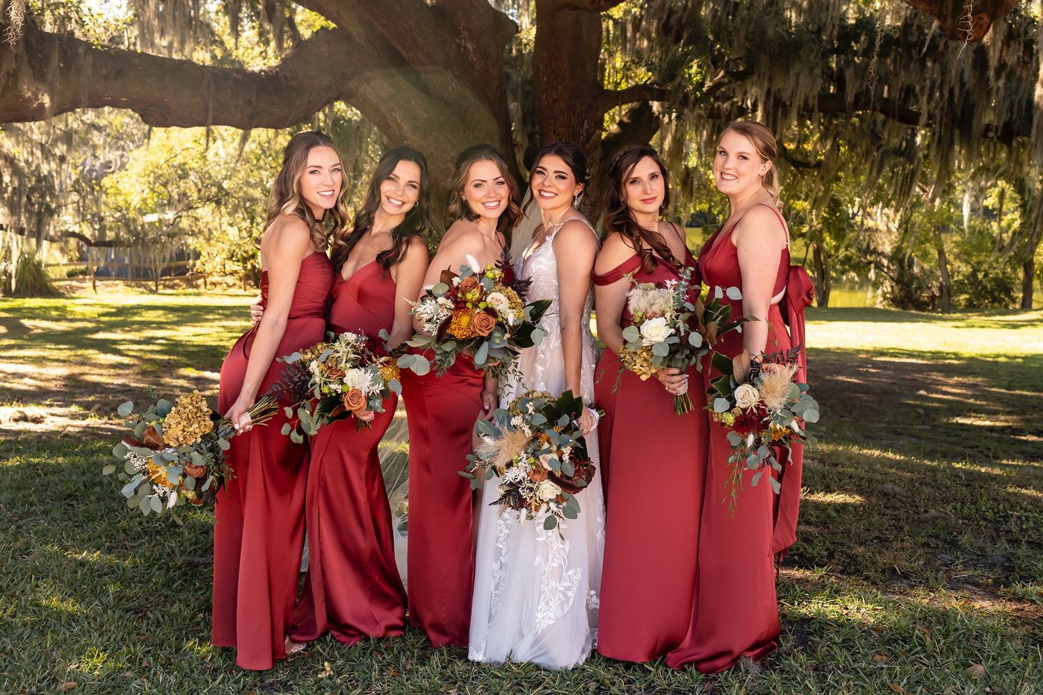 Bridesmaids in burgundy dresses and a bride in white pose with colorful floral bouquets in an outdoor garden setting.