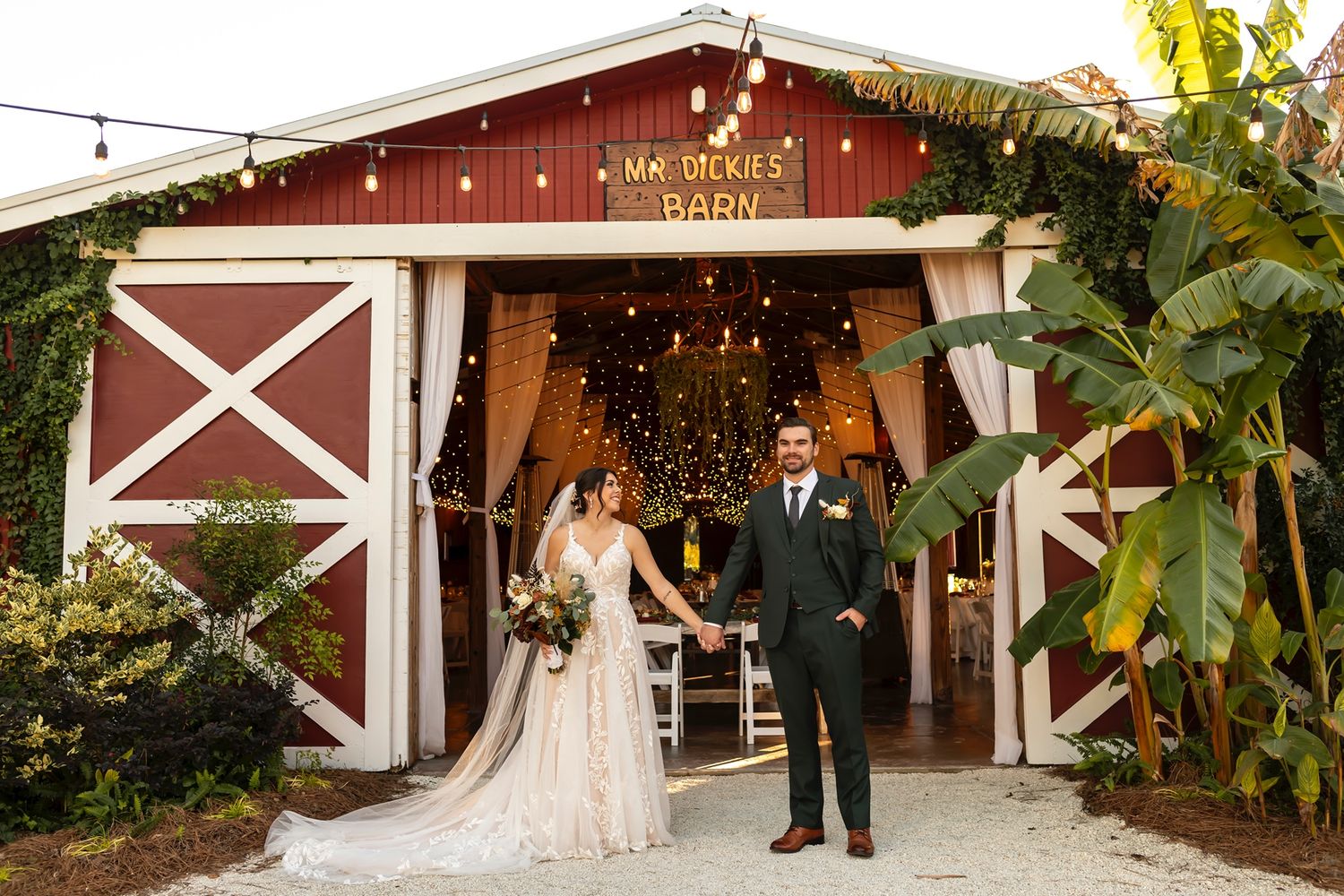 A couple stands outside a rustic red barn wedding venue with large wooden doors and twinkle lights glowing inside.