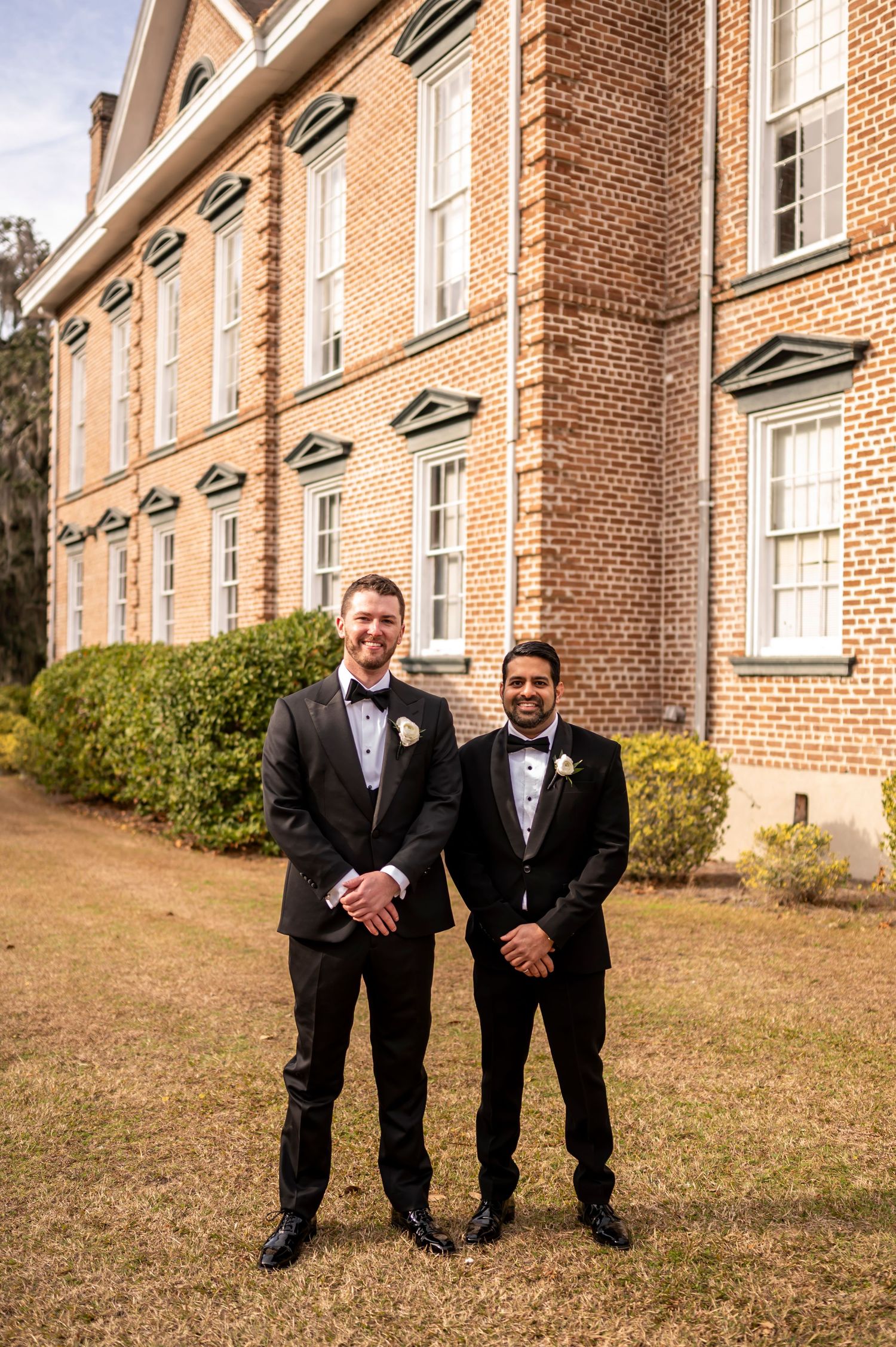 Two individuals in formal black tuxedos pose together on a lawn in front of a historic brick building.