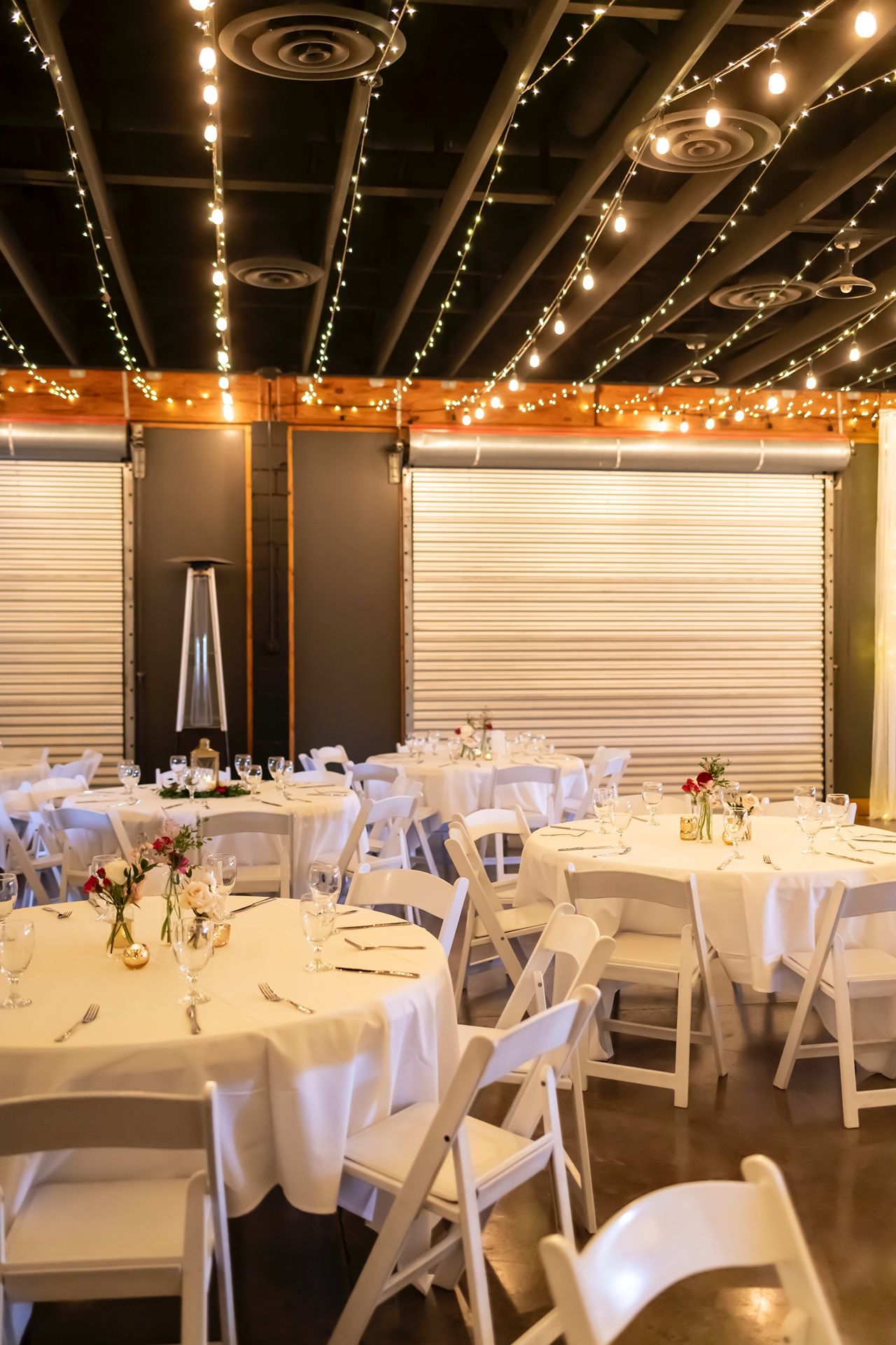 Indoor wedding reception venue decorated with string lights and white tables featuring folding chairs and elegant place settings.
