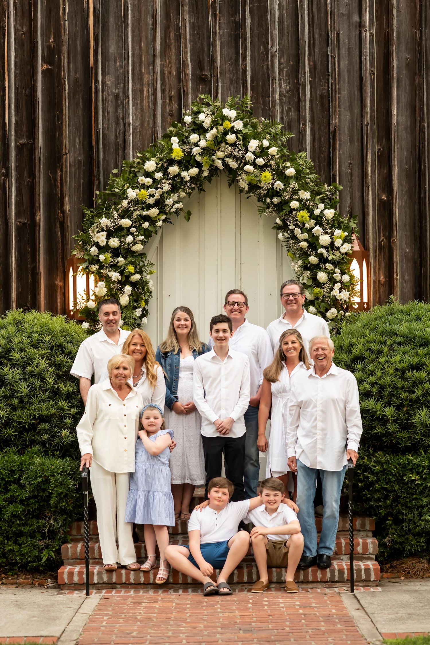 Multi-generational group poses together under beautiful floral arch with white roses against rustic wooden backdrop.