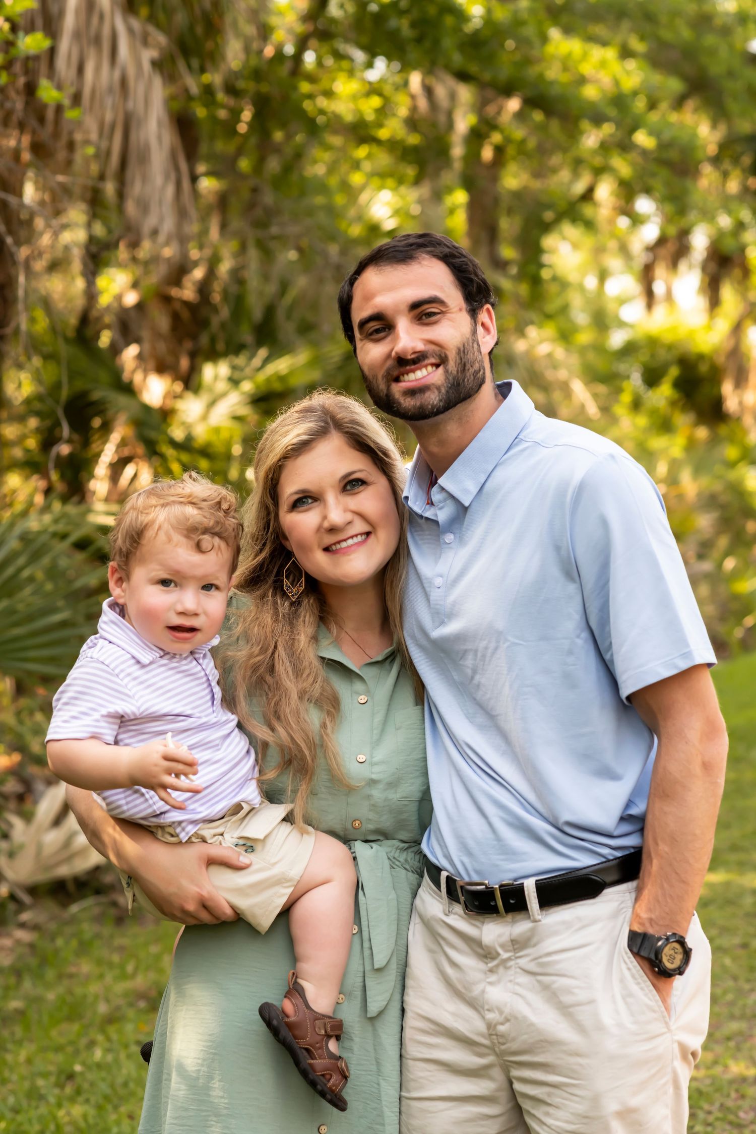 A family poses together outdoors in coordinated pastel outfits against a sunlit green backdrop.
