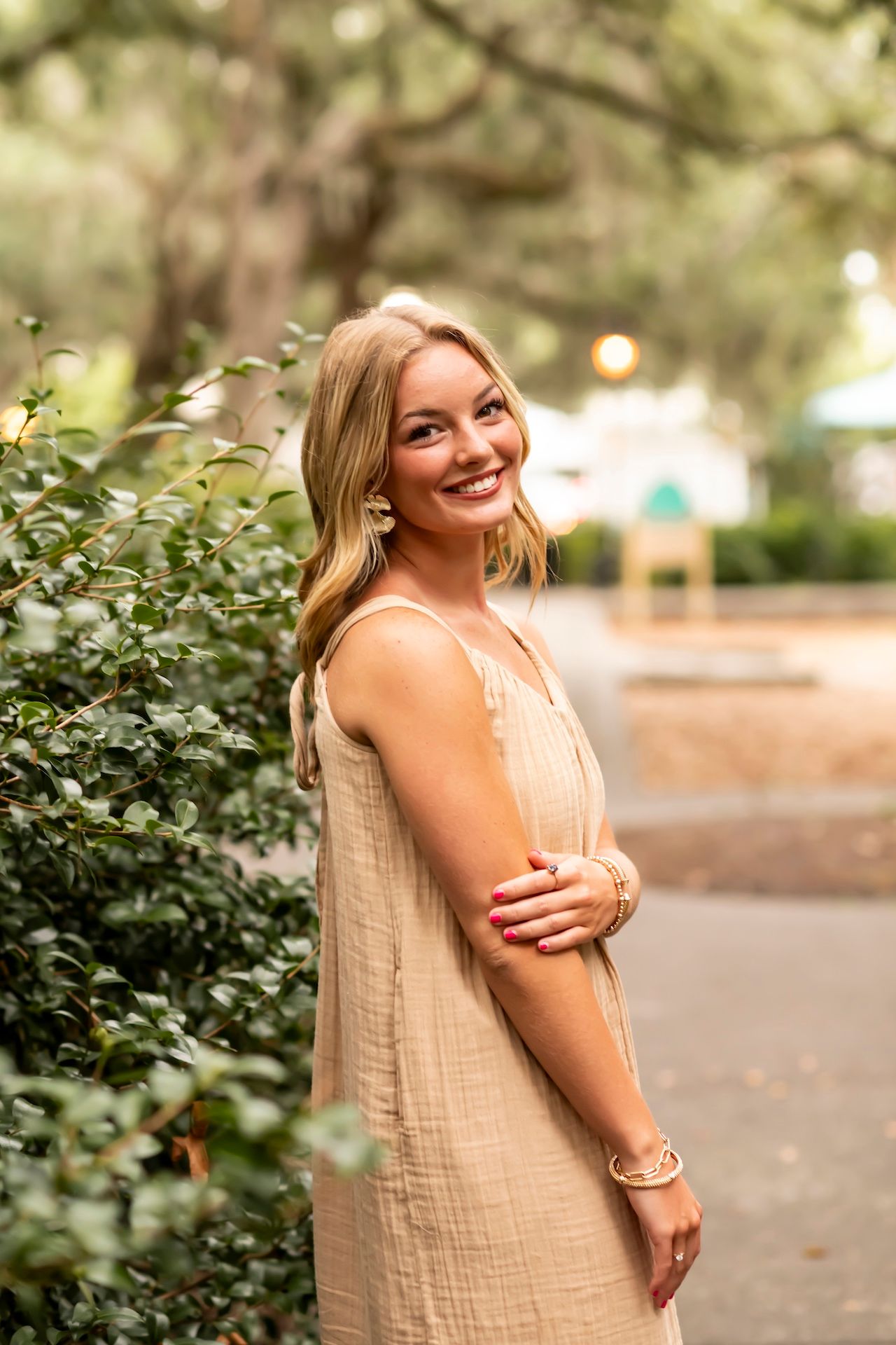 Person in beige sundress smiles brightly while posing outdoors near foliage.