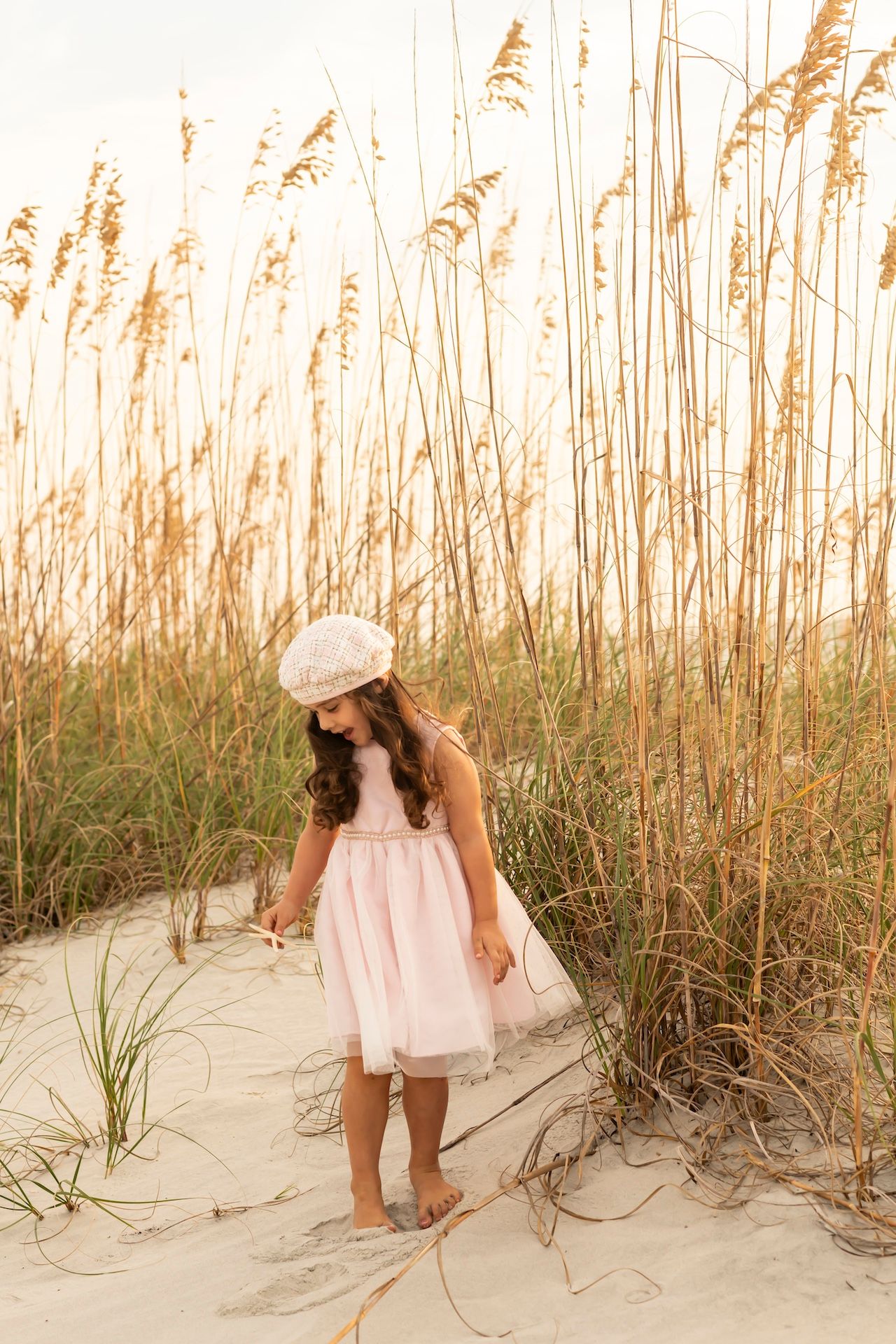 Young child in a pink dress and hat exploring sandy dunes with tall sea oats in warm sunset lighting.