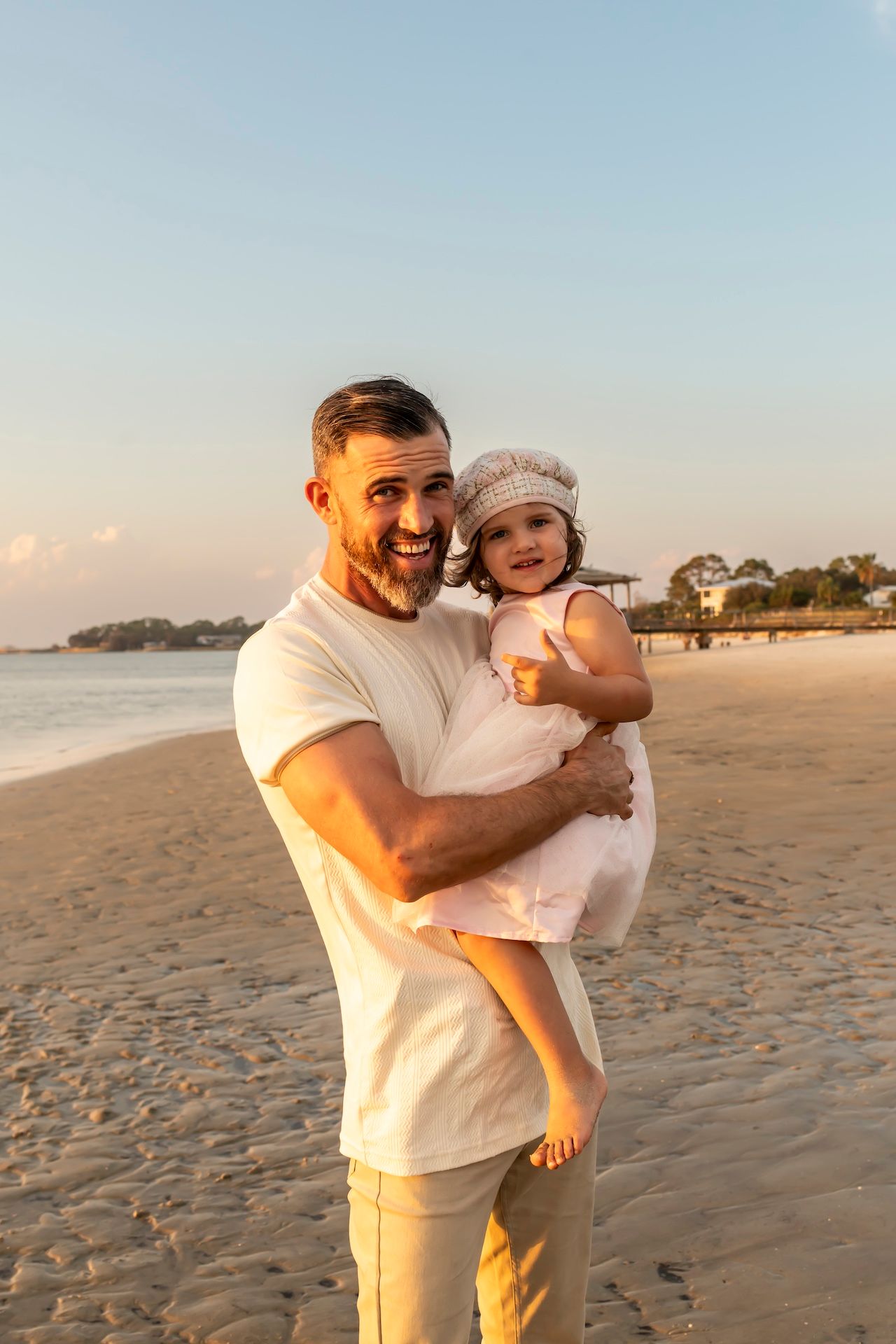 Warm sunset light illuminates a sandy beach as a person in a white t-shirt holds a small child wearing a knit hat.