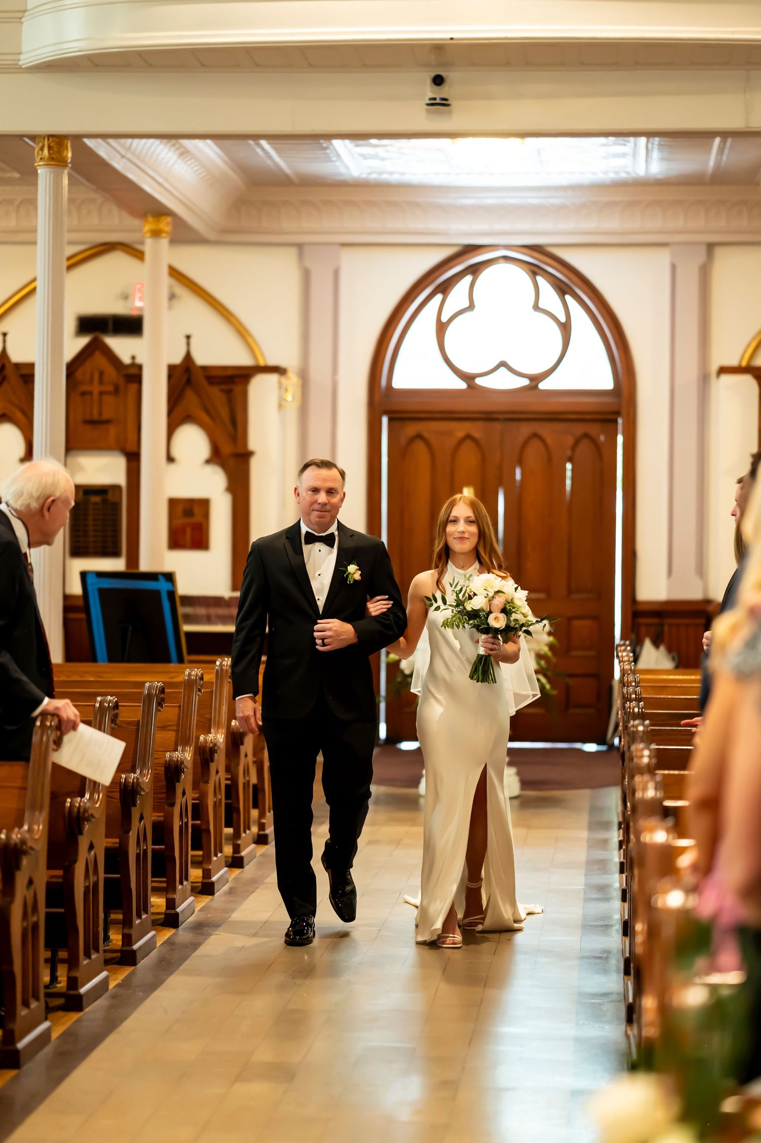 A couple walks down a church aisle with wooden pews and gothic architectural details during a solemn ceremony.