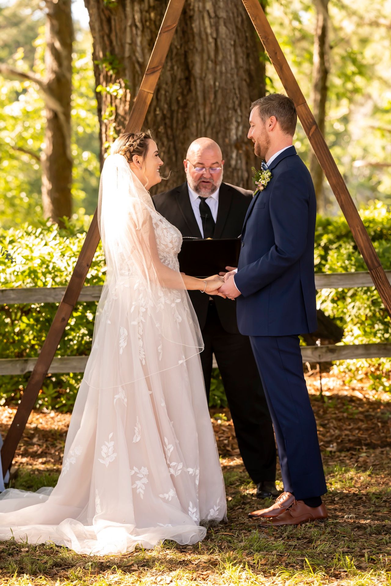 A wedding ceremony takes place under a wooden triangle arch in a sunlit forest setting, with the couple holding hands.