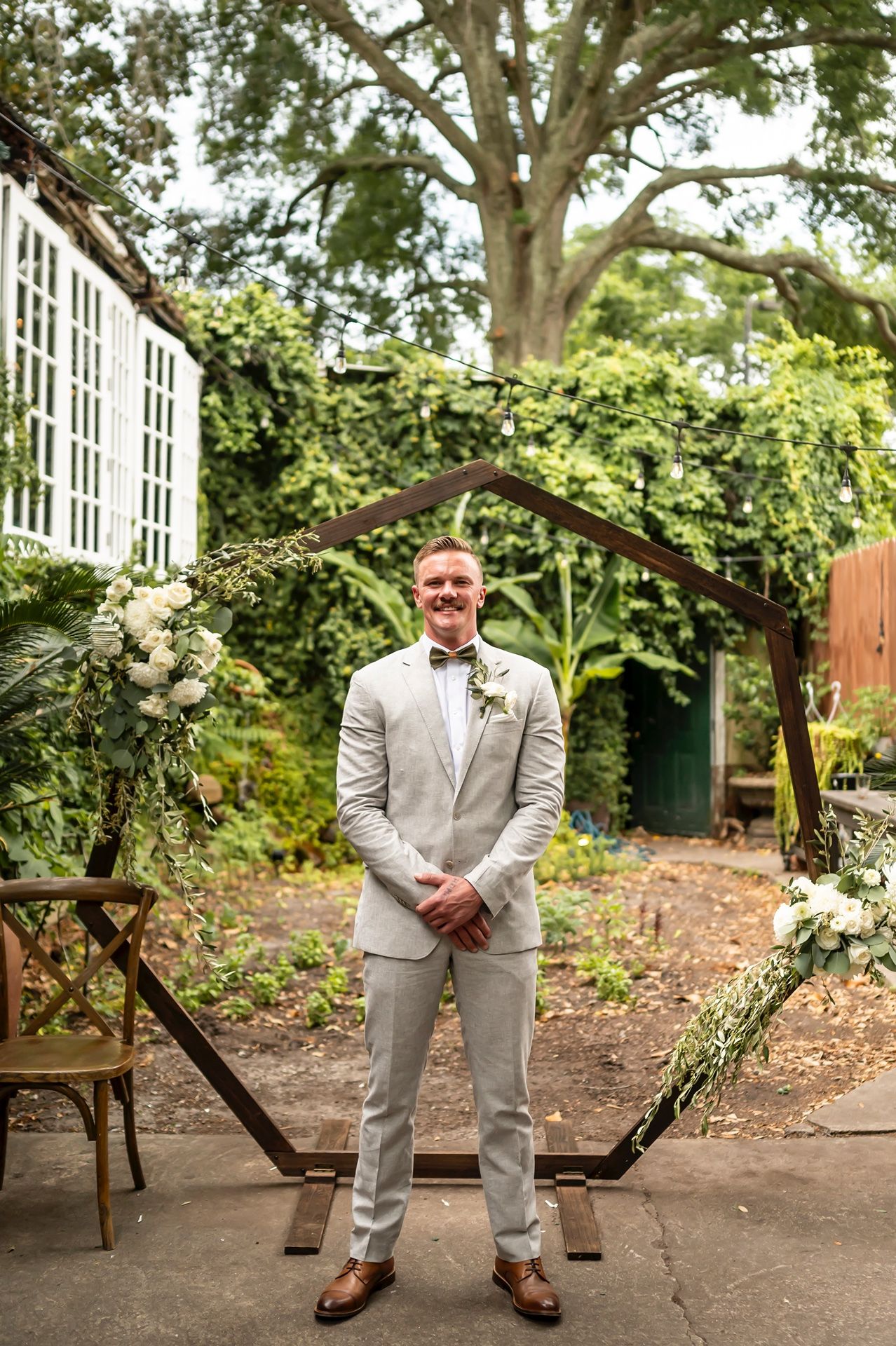 A person in a light gray suit and bowtie stands under a wooden arch decorated with white flowers in a garden setting.