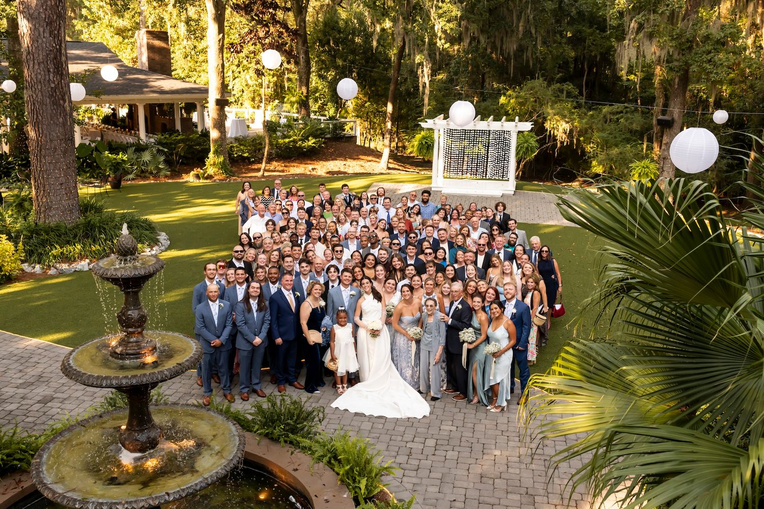 Large wedding group photo captured in front of a garden fountain with tropical landscaping and white decorative elements.