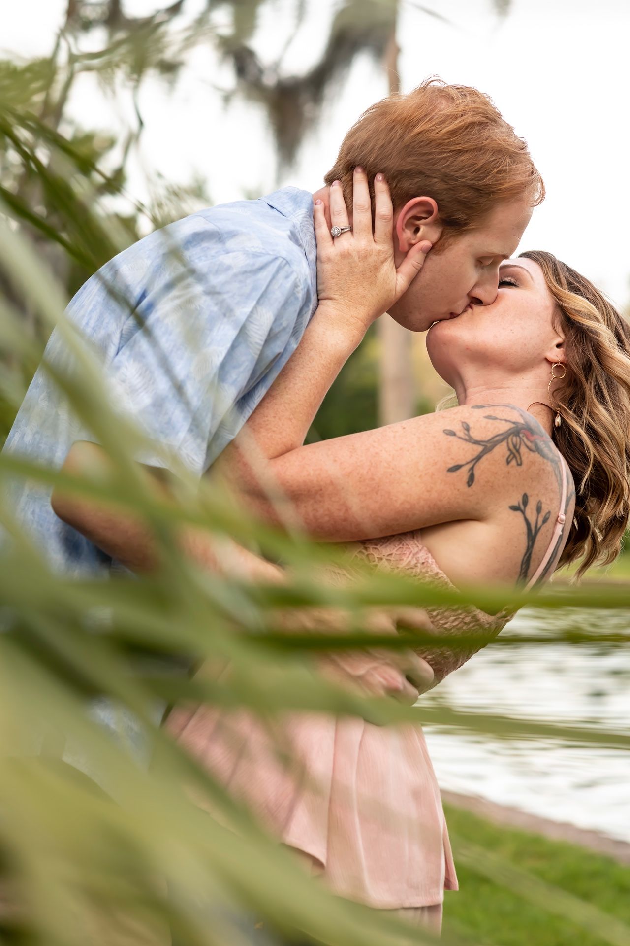 Romantic couple sharing a tender kiss outdoors by the water, framed by green foliage in soft natural lighting.