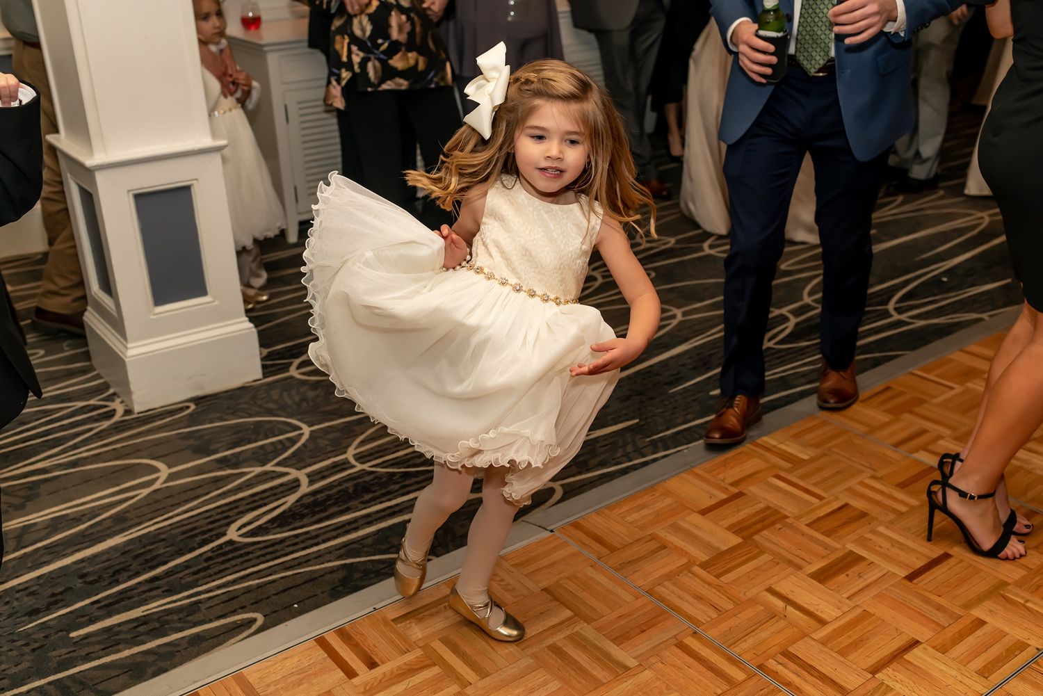 A young flower girl in a cream-colored dress twirls on a parquet dance floor during a wedding reception.