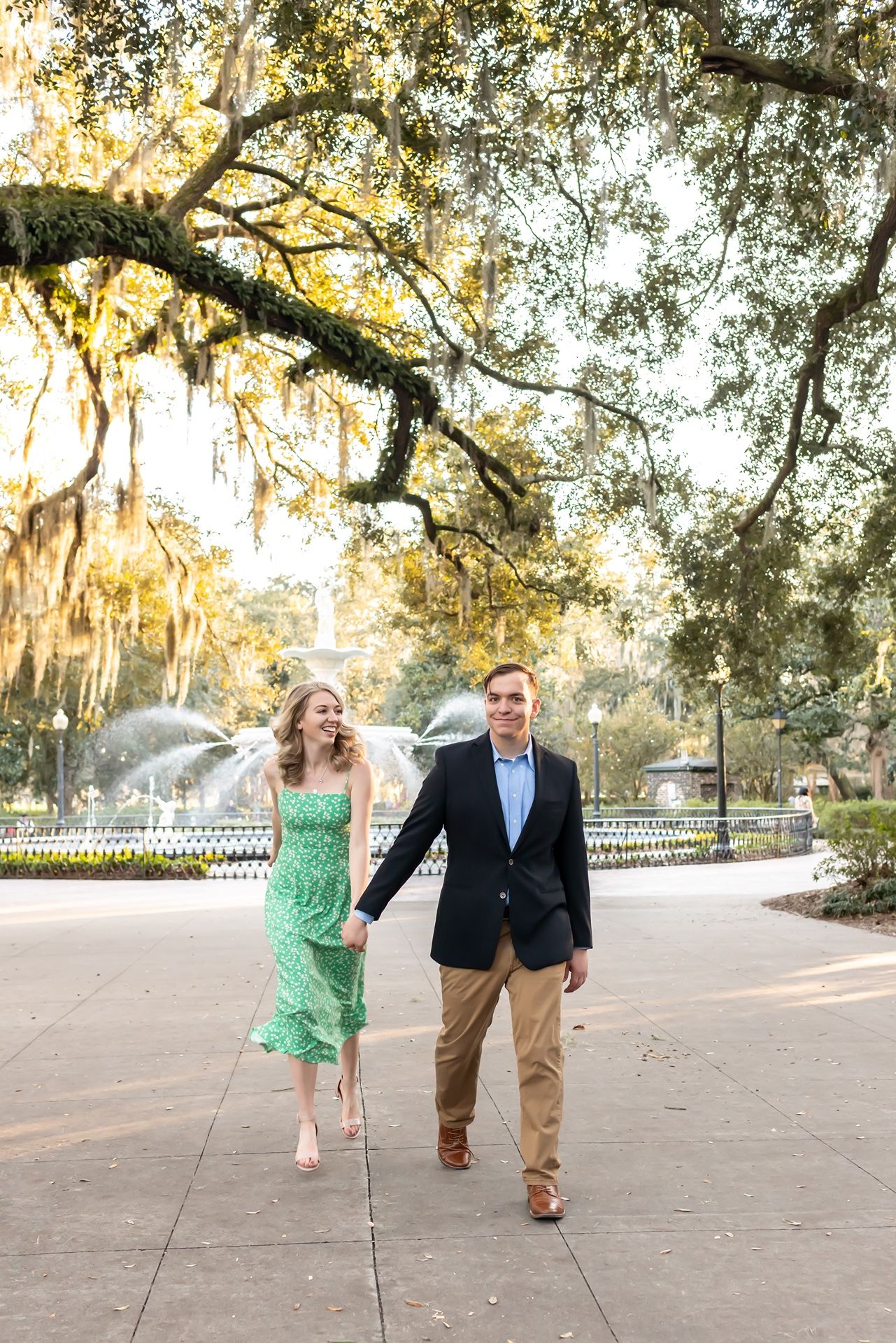 A couple strolls beneath Spanish moss draped oak trees in a romantic Southern park setting at sunset.