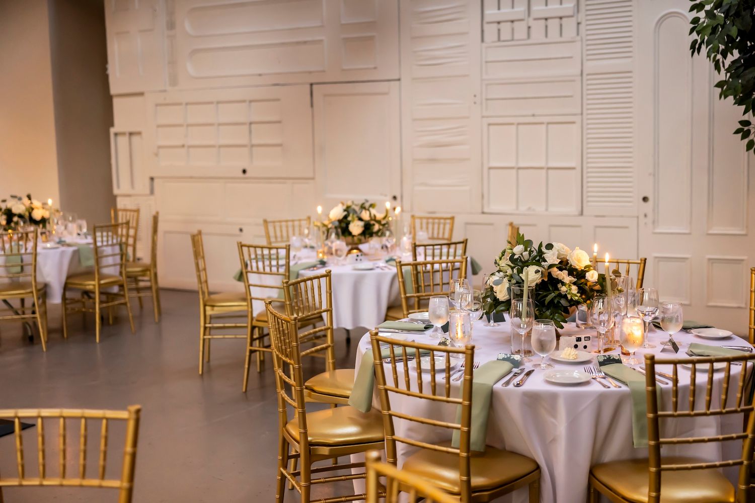 Elegant wedding reception venue features round tables with white linens, gold chiavari chairs, and soft lighting.