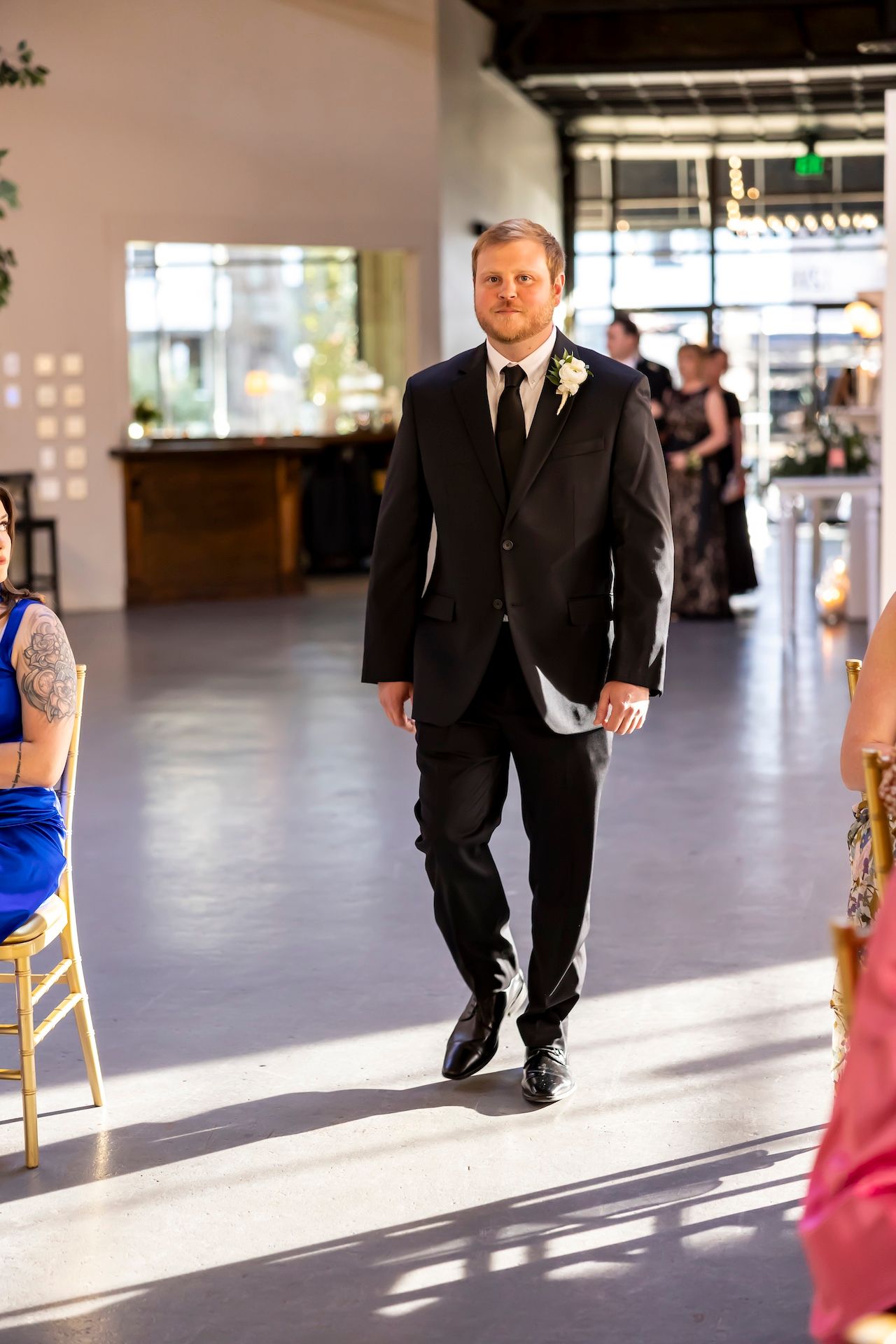 A groom in black suit walking down a sunlit indoor hallway with wooden chairs lining the sides.