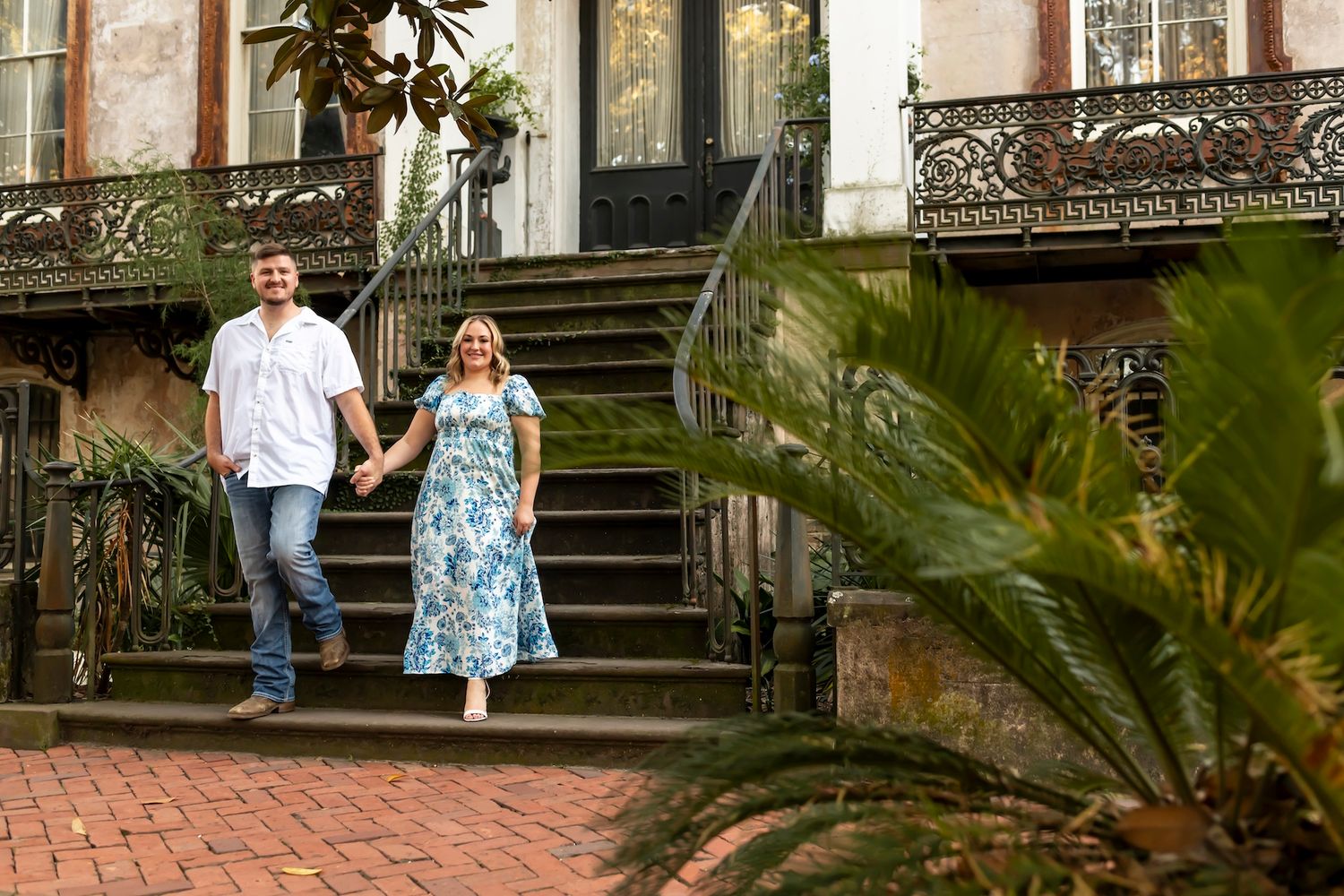 A couple descends ornate brick stairs outside a historic building with wrought iron railings and tropical plants.
