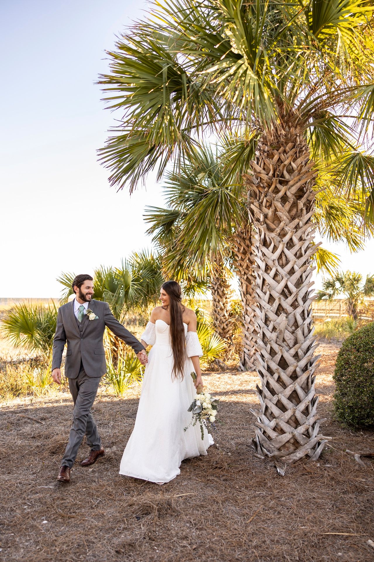 A couple shares joyful moments among palm trees on a beach during their outdoor wedding celebration at sunset.