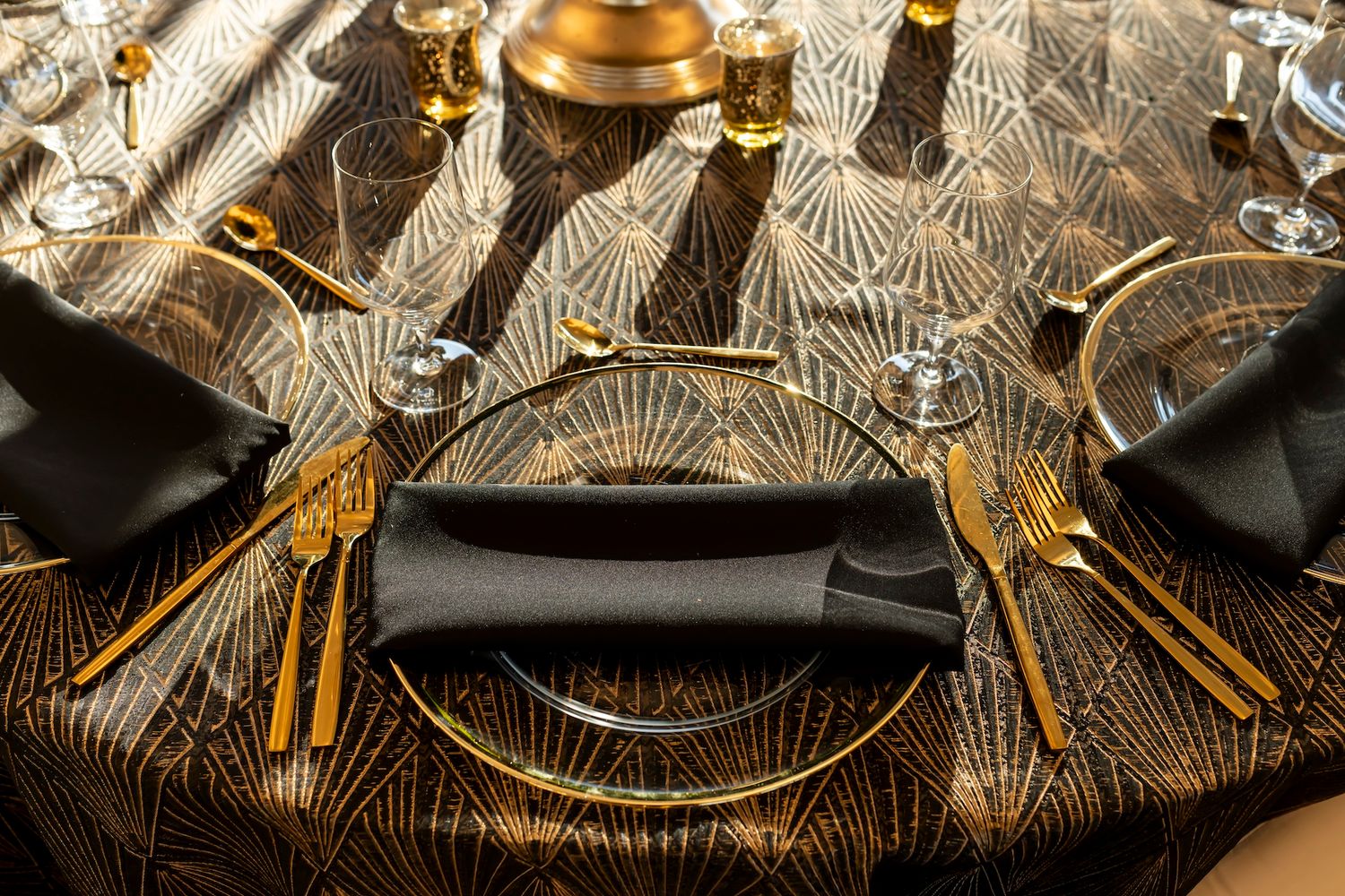 Elegant table setting with gold cutlery and black napkins on a textured glass charger plate with art deco pattern.