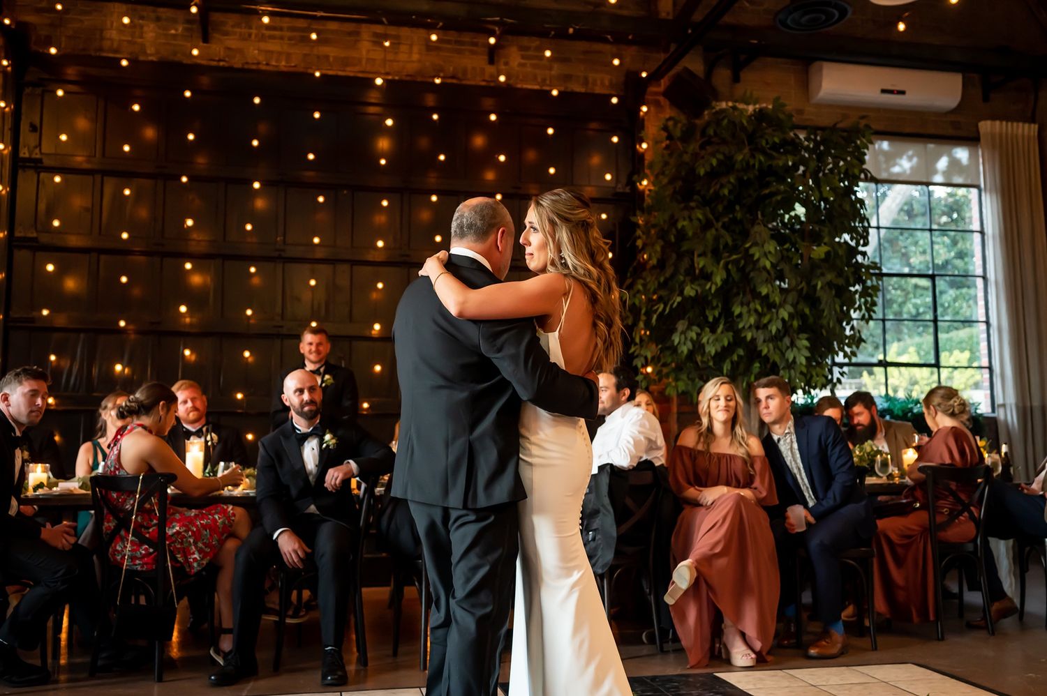 Couple shares their first dance as guests look on during an evening wedding reception with twinkling lights overhead.