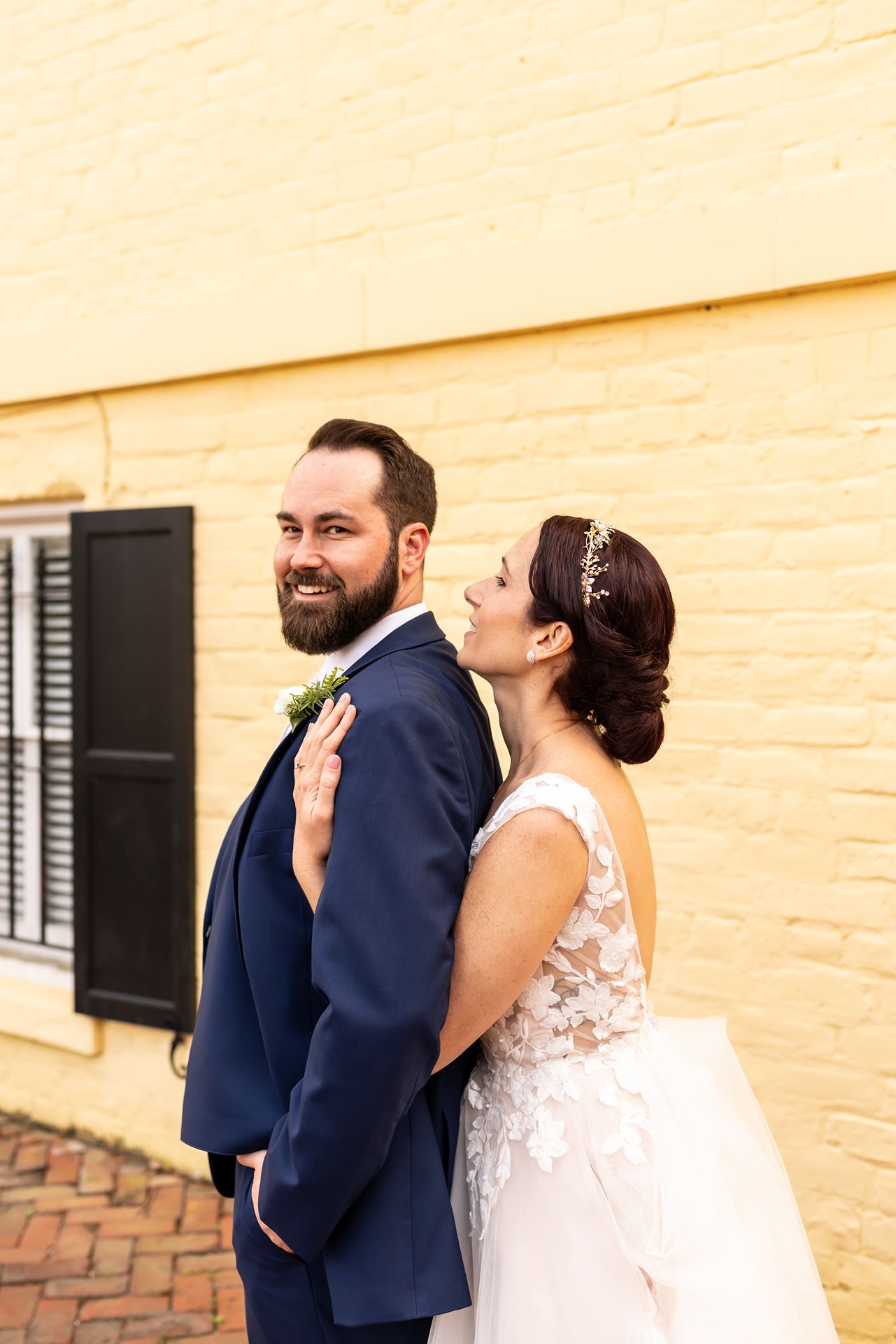 A couple in wedding attire pose together in front of a yellow brick wall on their special day.