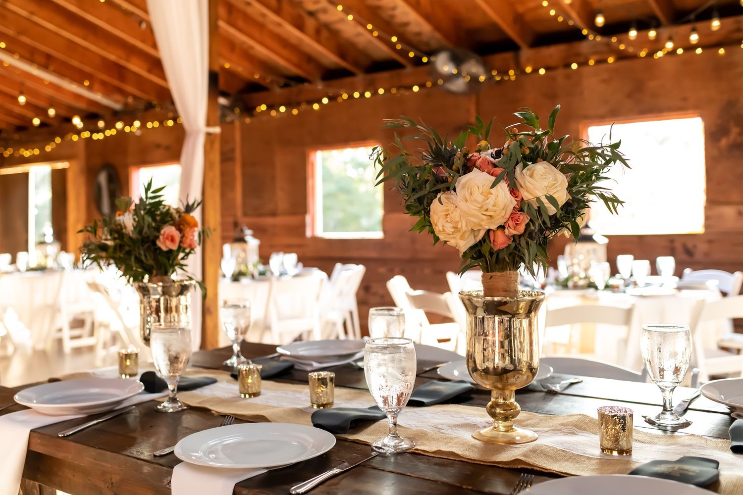Elegant rustic wedding reception venue with wooden beams, string lights, and floral centerpieces on white tablescapes.