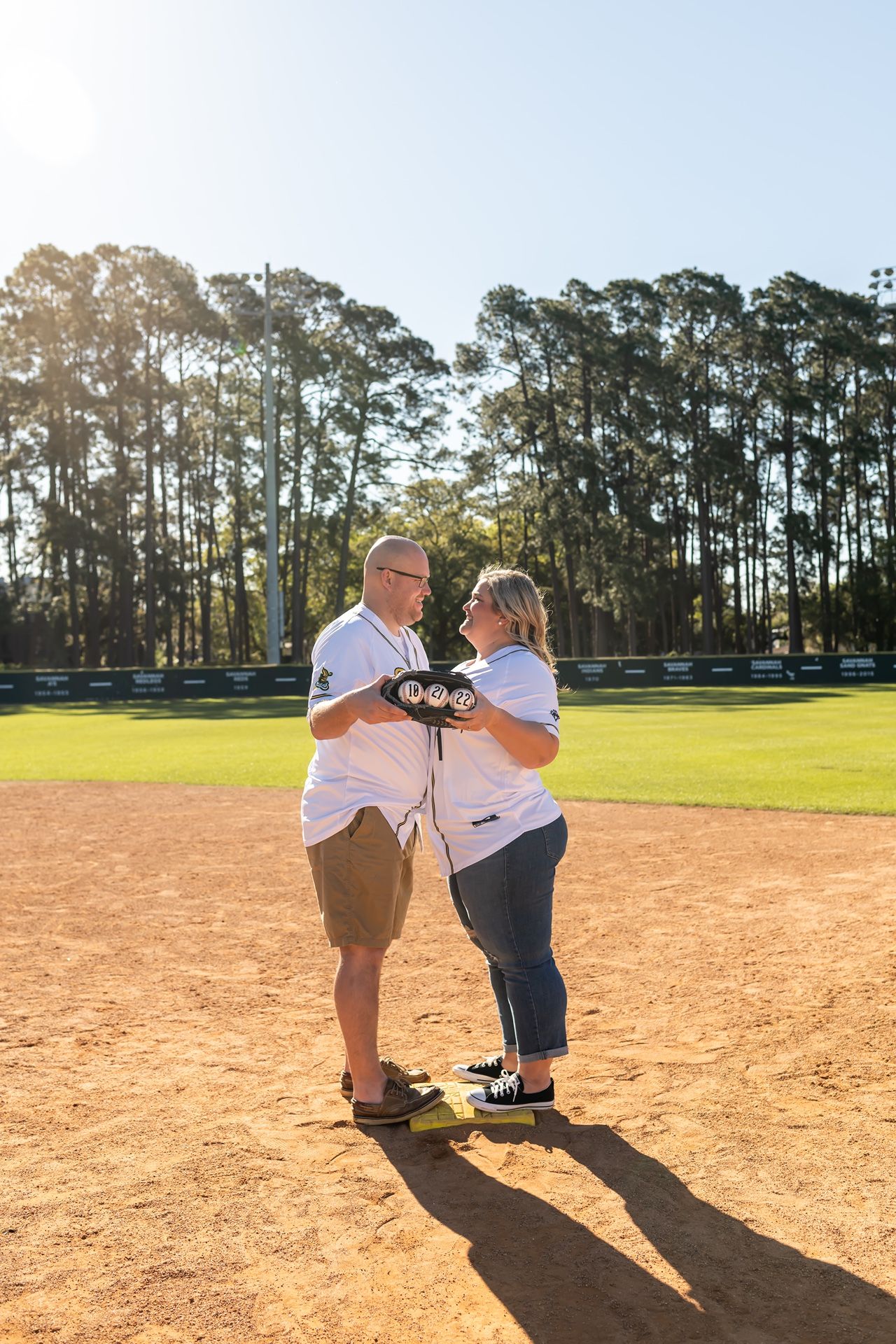 Two people interact playfully on a sunlit dirt baseball field with tall pine trees in the background during golden hour.
