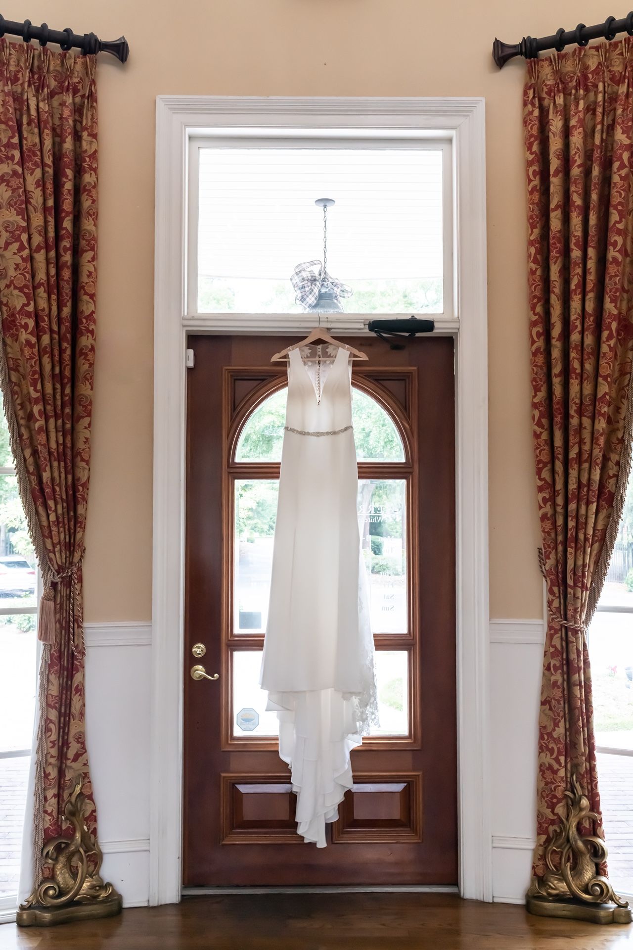 A white wedding dress hangs elegantly against a wooden front door framed by ornate floral curtains in a stately entryway.