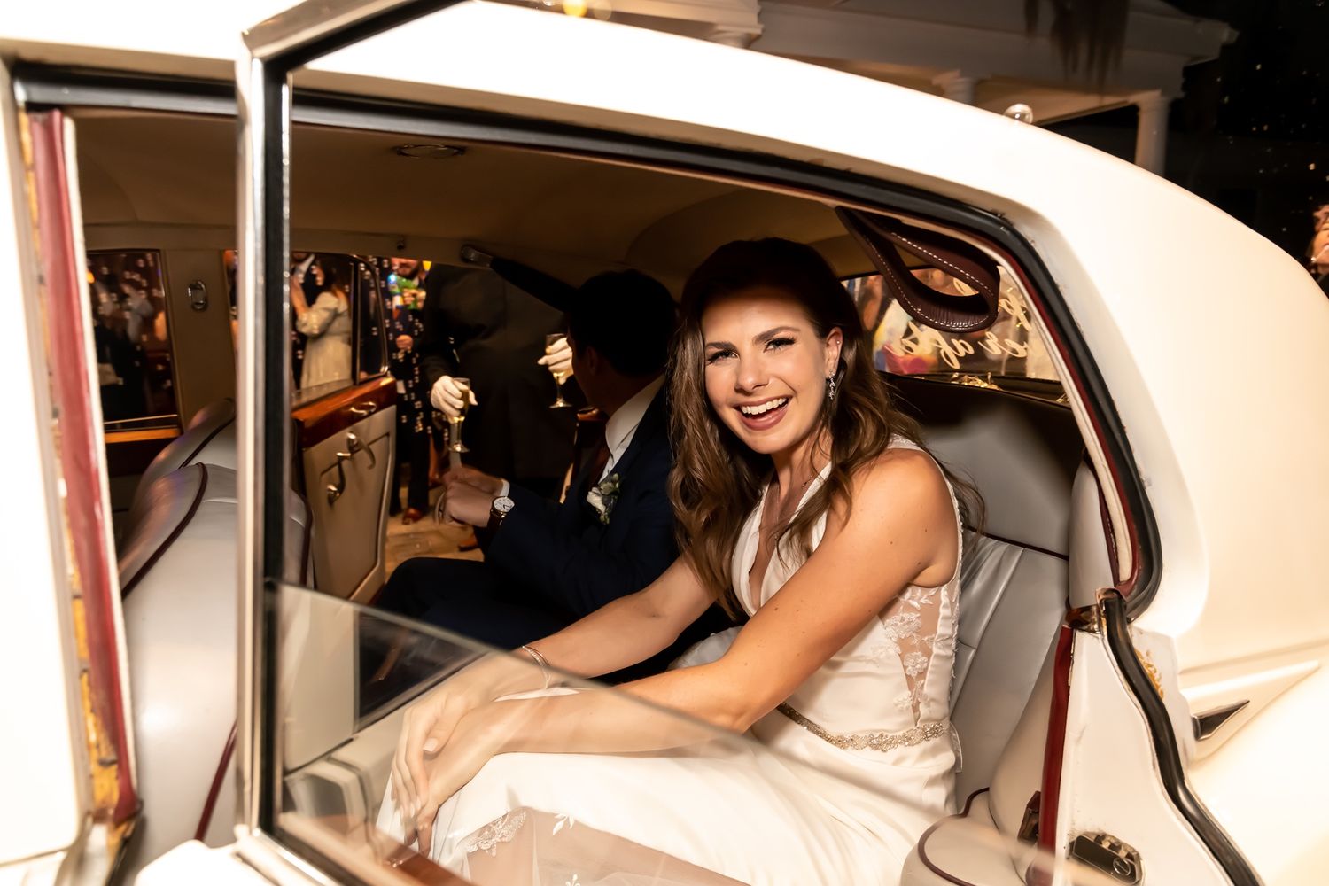 Someone wearing an elegant white dress sits smiling in the back seat of a vintage cream-colored car.
