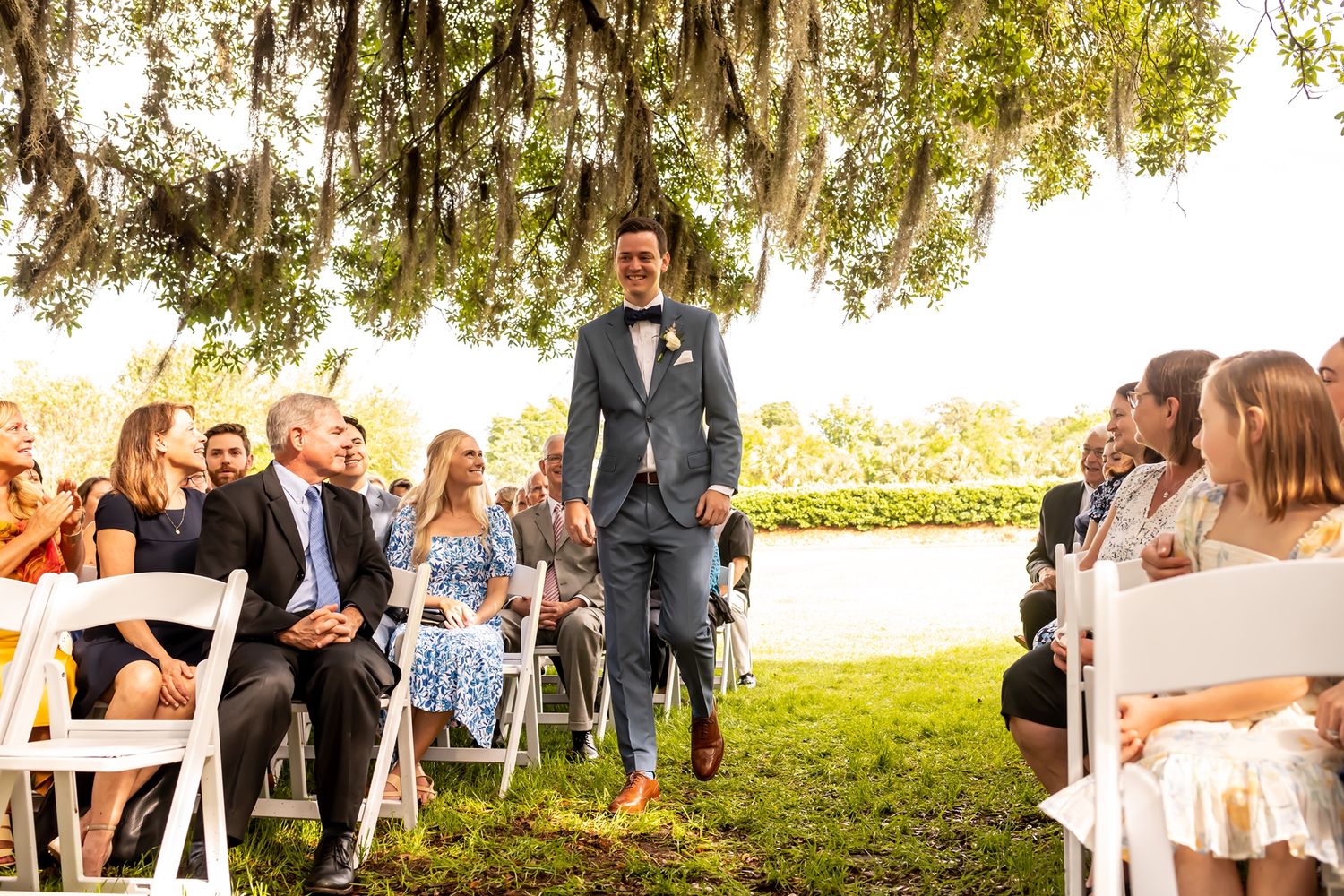 Wedding guests seated in white chairs at an outdoor ceremony smiling under moss-draped trees on a sunny afternoon.