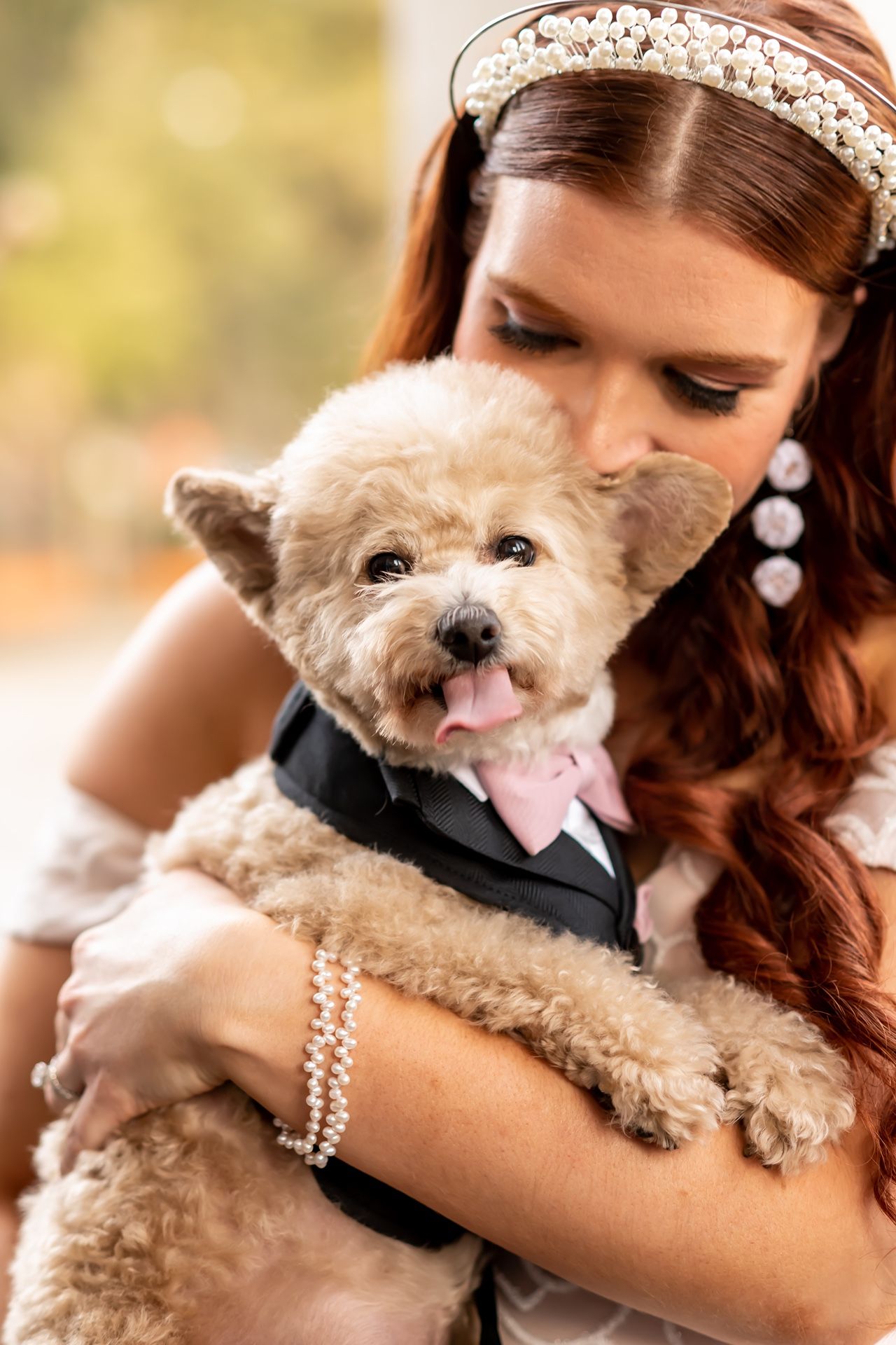 Smiling fluffy dog wearing a tuxedo being held by someone in a bridal dress with pearl accessories in warm outdoor lighting.