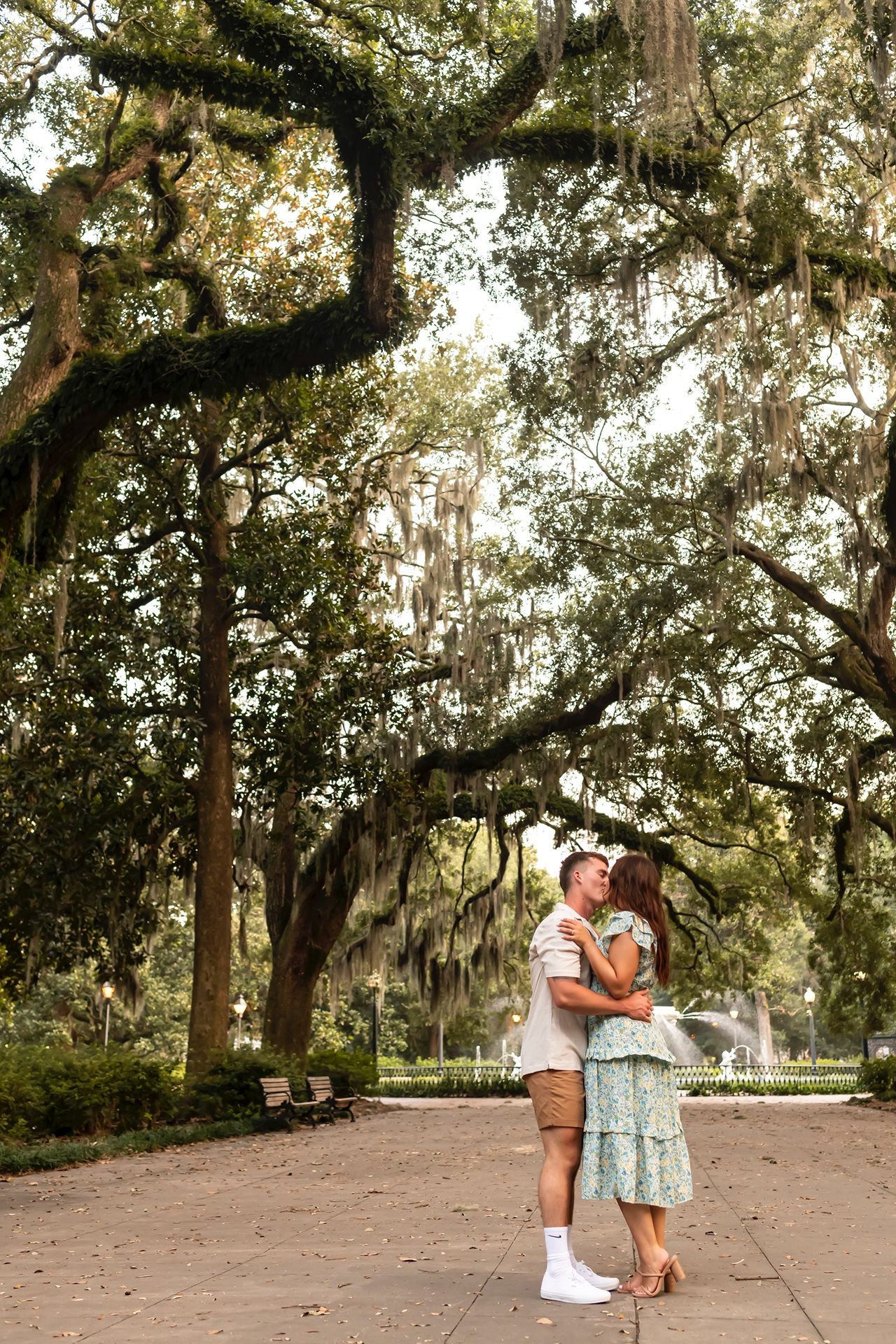 A couple embraces on a path beneath towering oak trees draped with Spanish moss in a romantic Southern setting.
