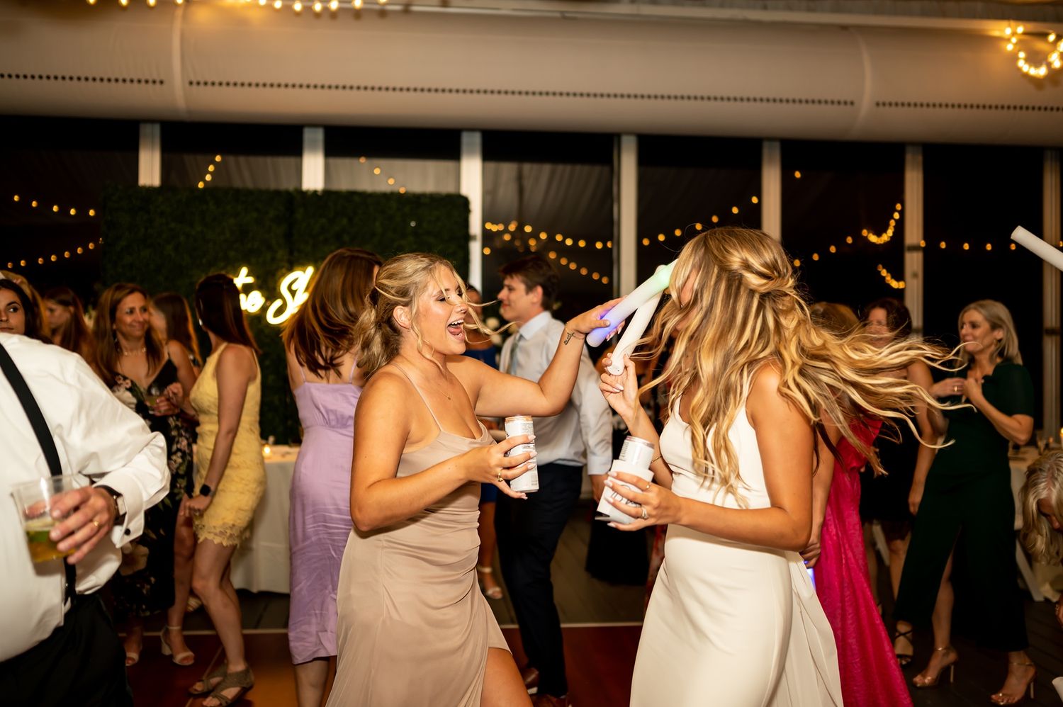 People dancing energetically at a wedding reception with festive string lights and greenery decorations in the background.