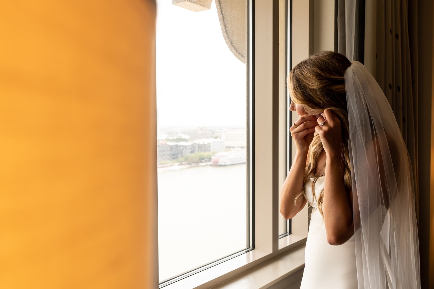 Person in white wedding dress and veil looking through window with orange walls, creating silhouette against natural light.