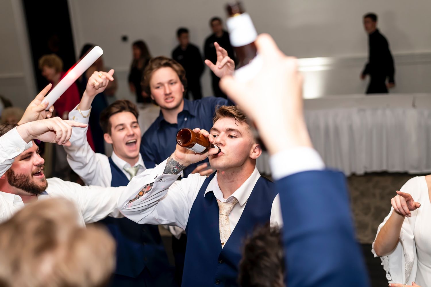 A group of party guests in blue suits celebrate wildly while one person chugs from a bottle at a formal event.