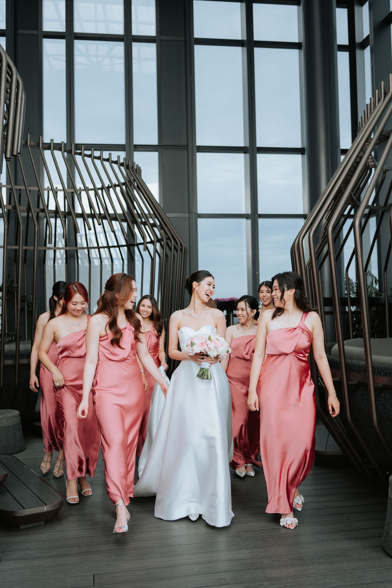 A wedding party in coordinated pink bridesmaid dresses and white bridal gown walking together in a modern indoor venue.