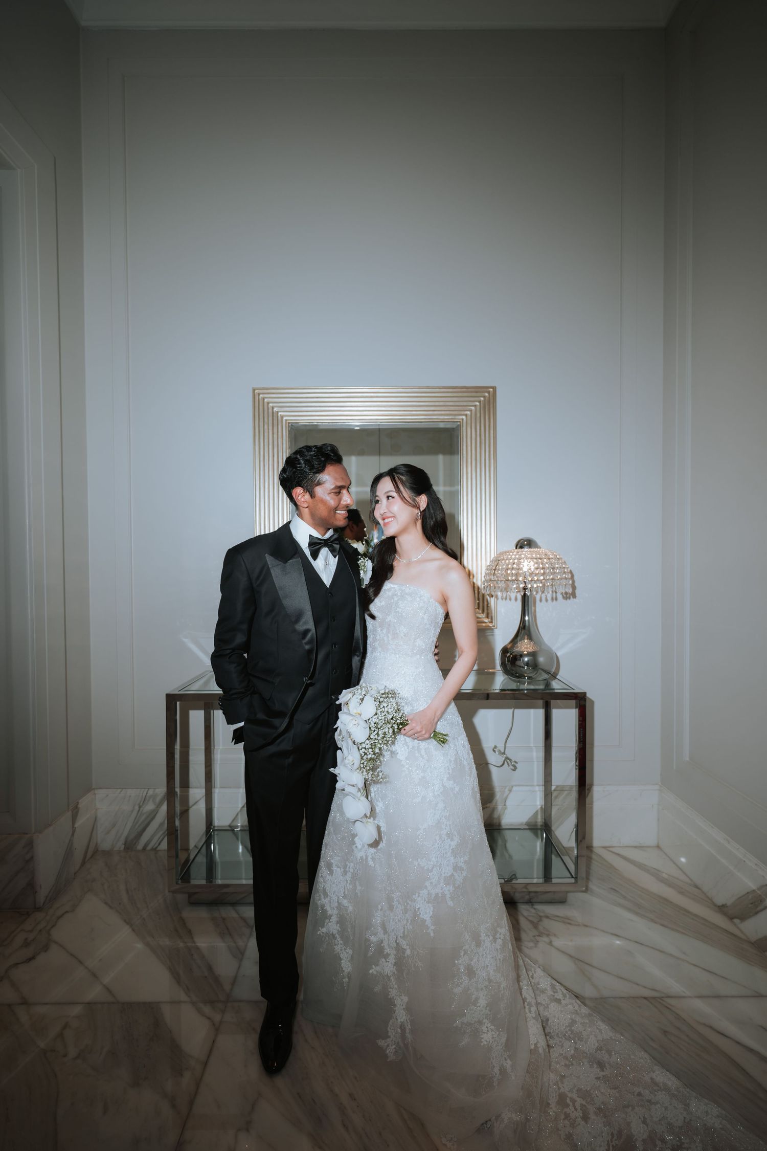 A bride and groom share romantic moments in an elegant white room with minimalist decor and soft lighting.