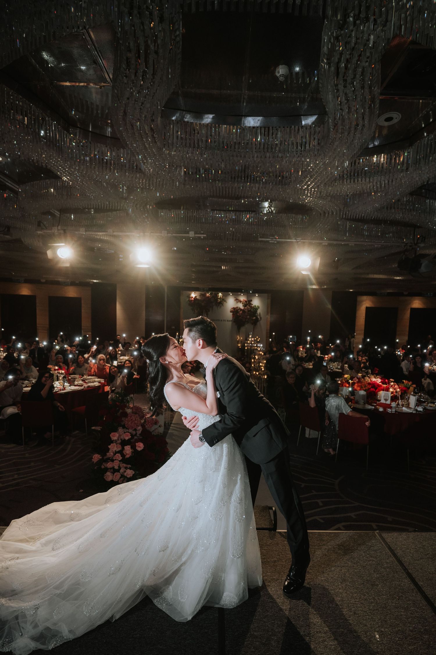 A couple shares a romantic dance moment on their wedding day in a dimly lit ballroom with spotlights overhead.