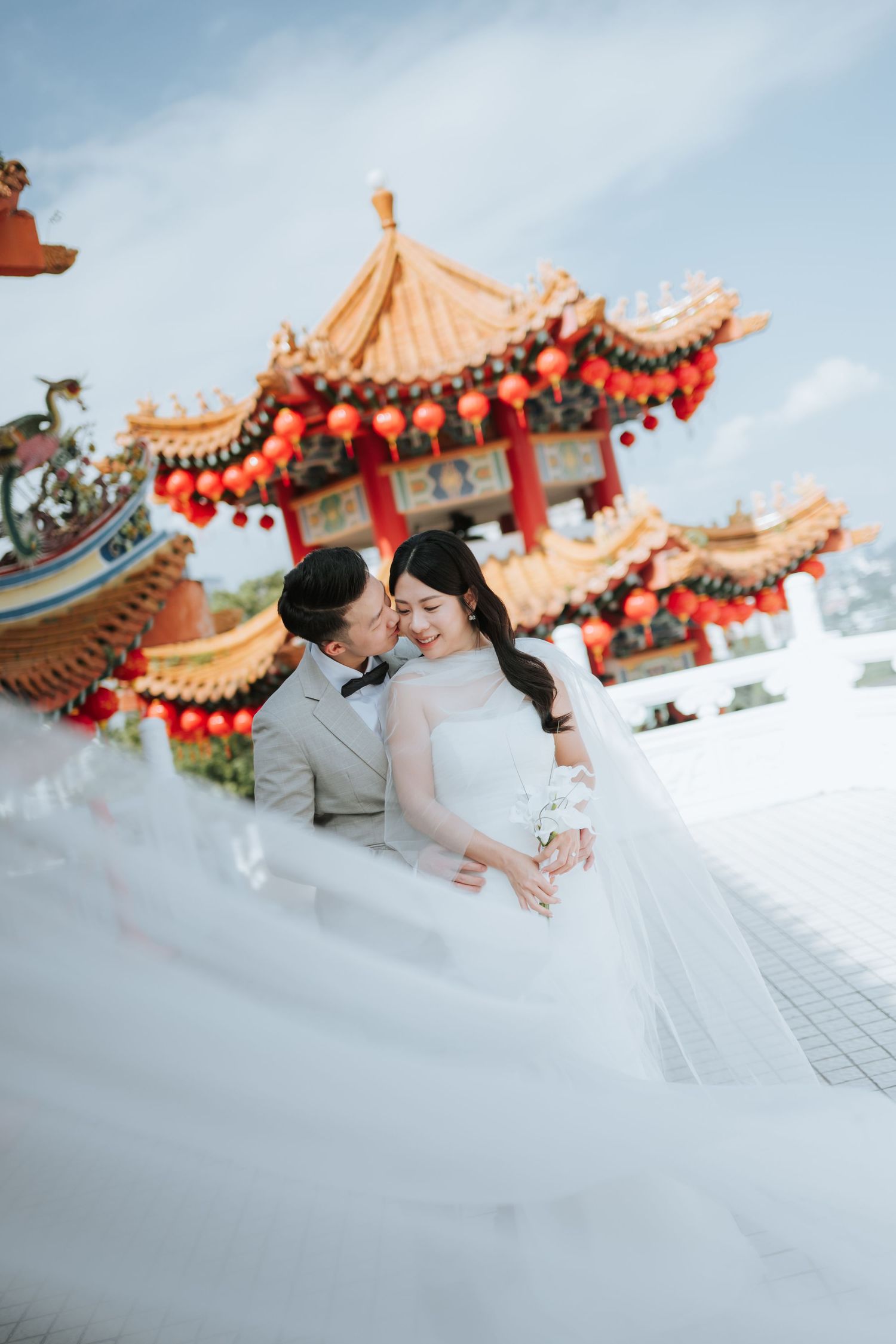 A romantic wedding photo captured in front of a traditional Chinese temple decorated with red lanterns and ornate details.