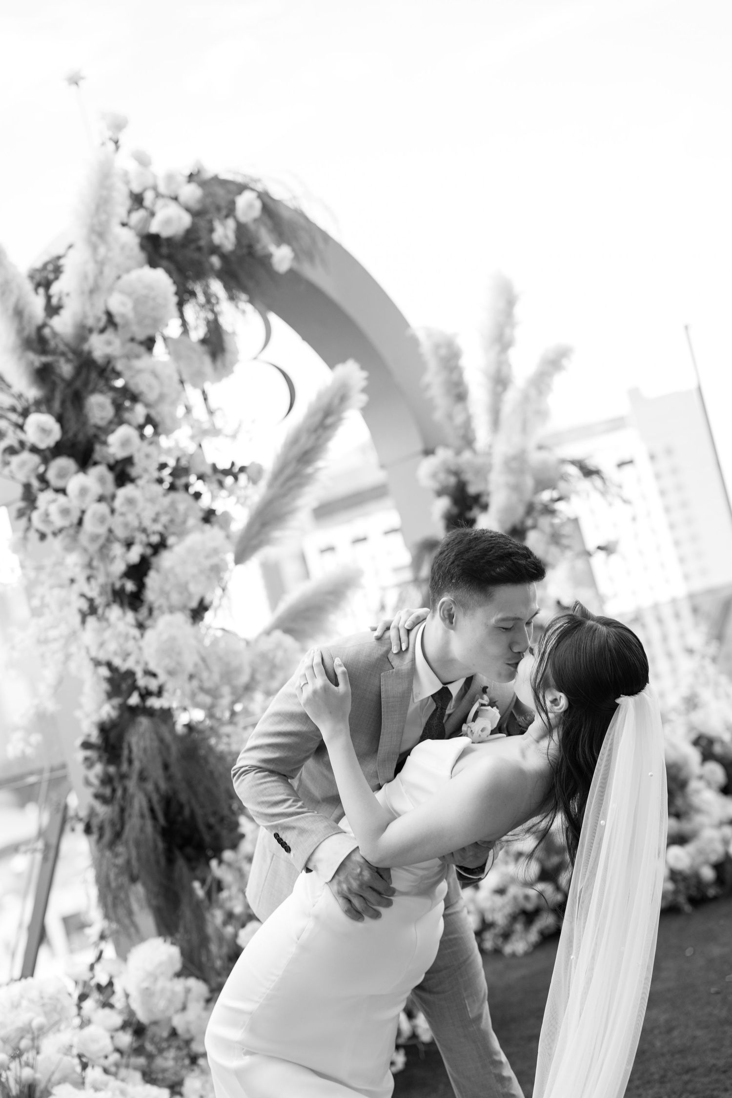 A romantic wedding moment captured in black and white beneath a floral arch during an outdoor ceremony.