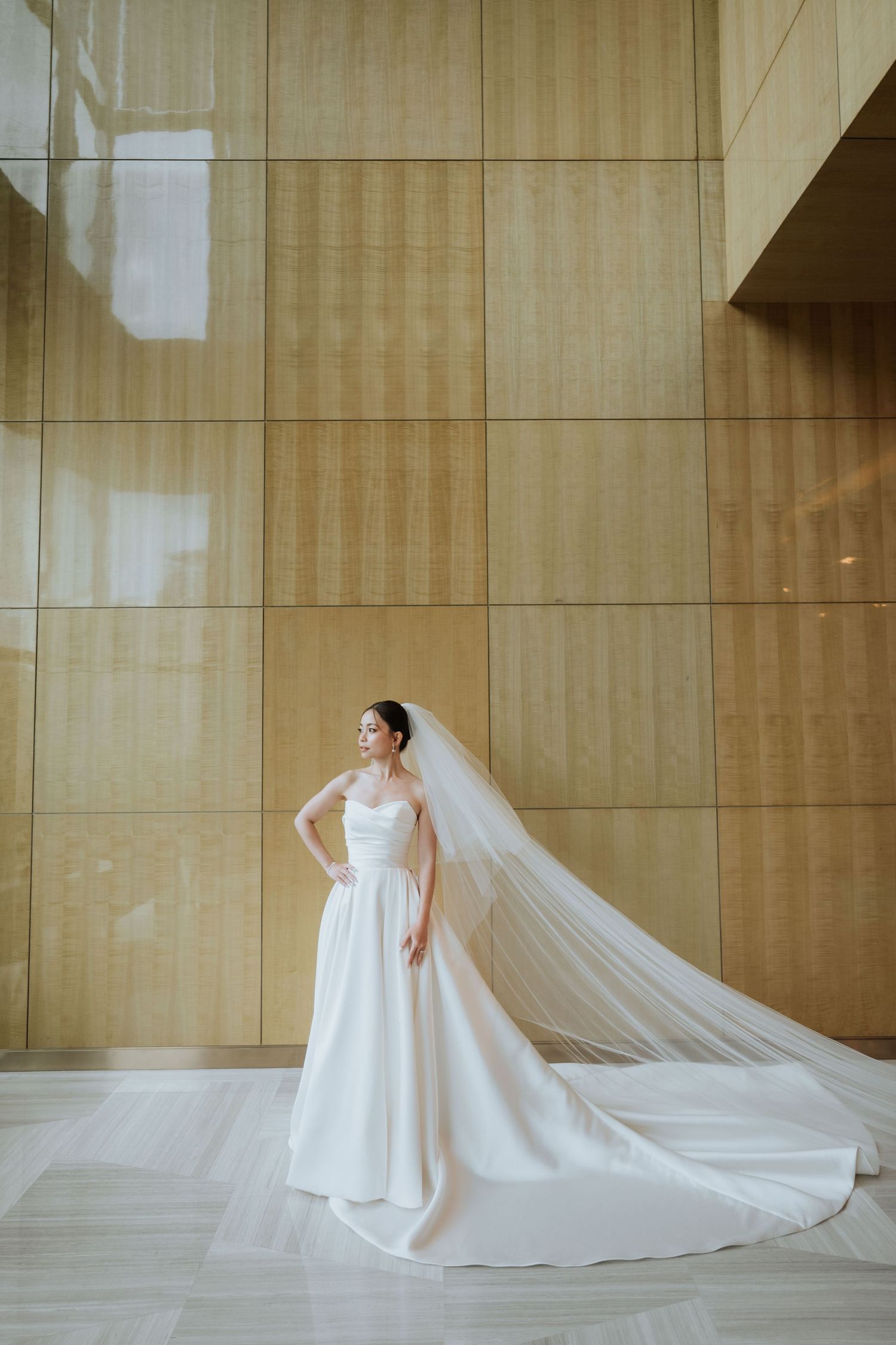 A bride in an elegant white wedding gown with a flowing cathedral-length veil stands against a modern gold-paneled wall.