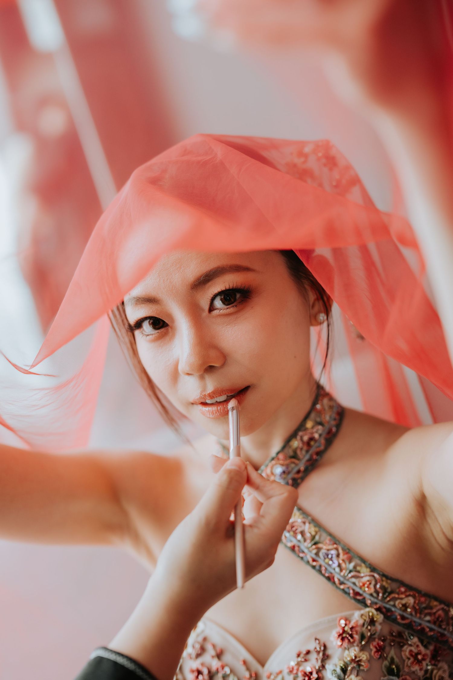 Portrait through pink veil with floral patterned dress and dramatic eye makeup in soft lighting.