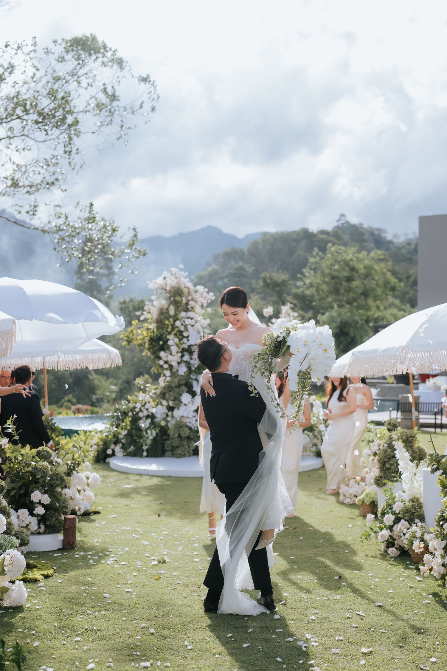 An elegant outdoor wedding ceremony with white umbrellas and floral arrangements against a mountain backdrop on a sunny day.