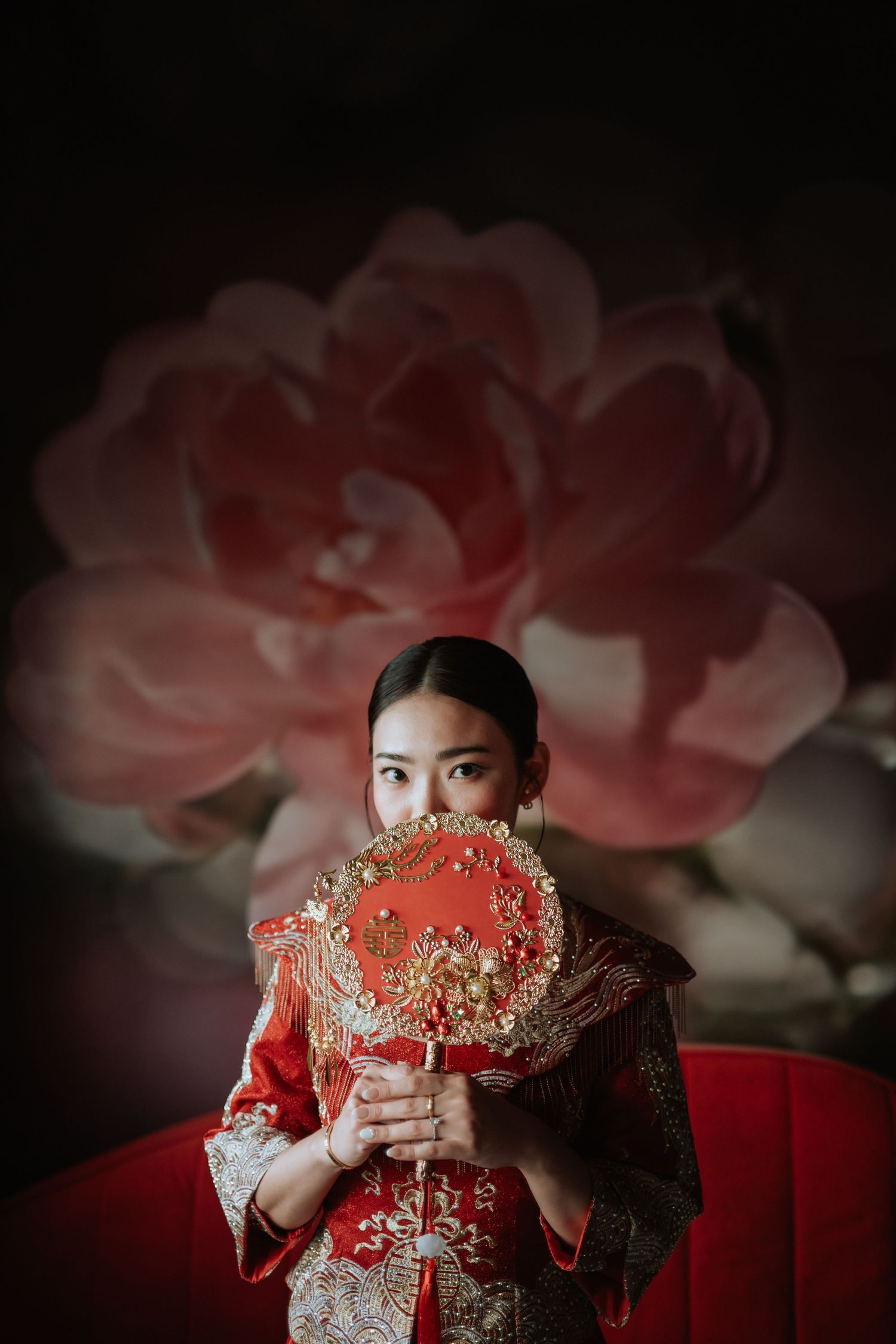 A bride in traditional red Chinese wedding attire holds an ornate fan against a backdrop of a large pink rose.