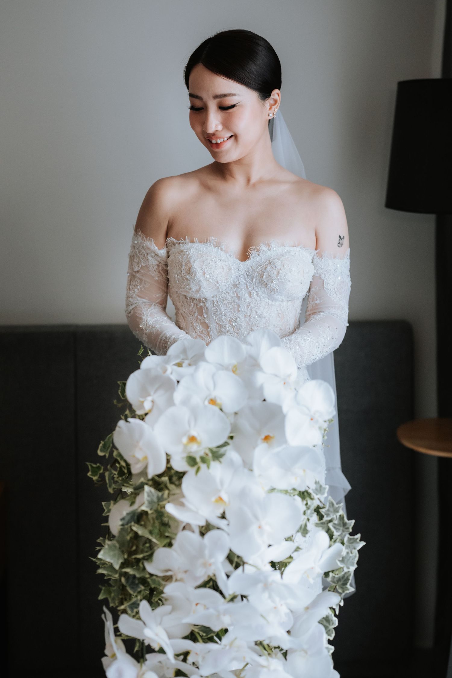 Bride in off-shoulder lace wedding dress holds elegant cascading white orchid bouquet with greenery accents.