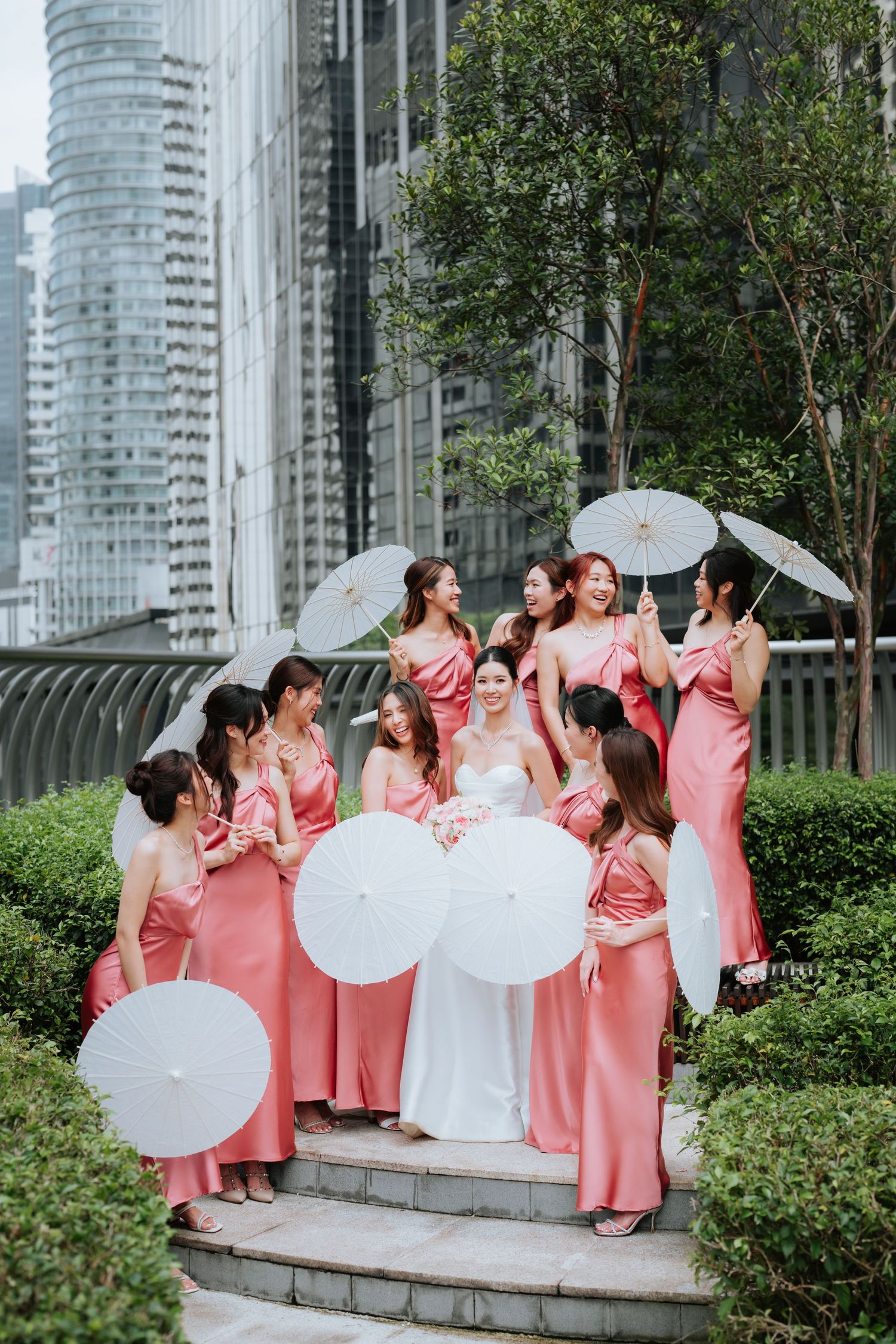 A bridal party wearing matching pink dresses poses on garden steps with white parasols against modern city buildings.