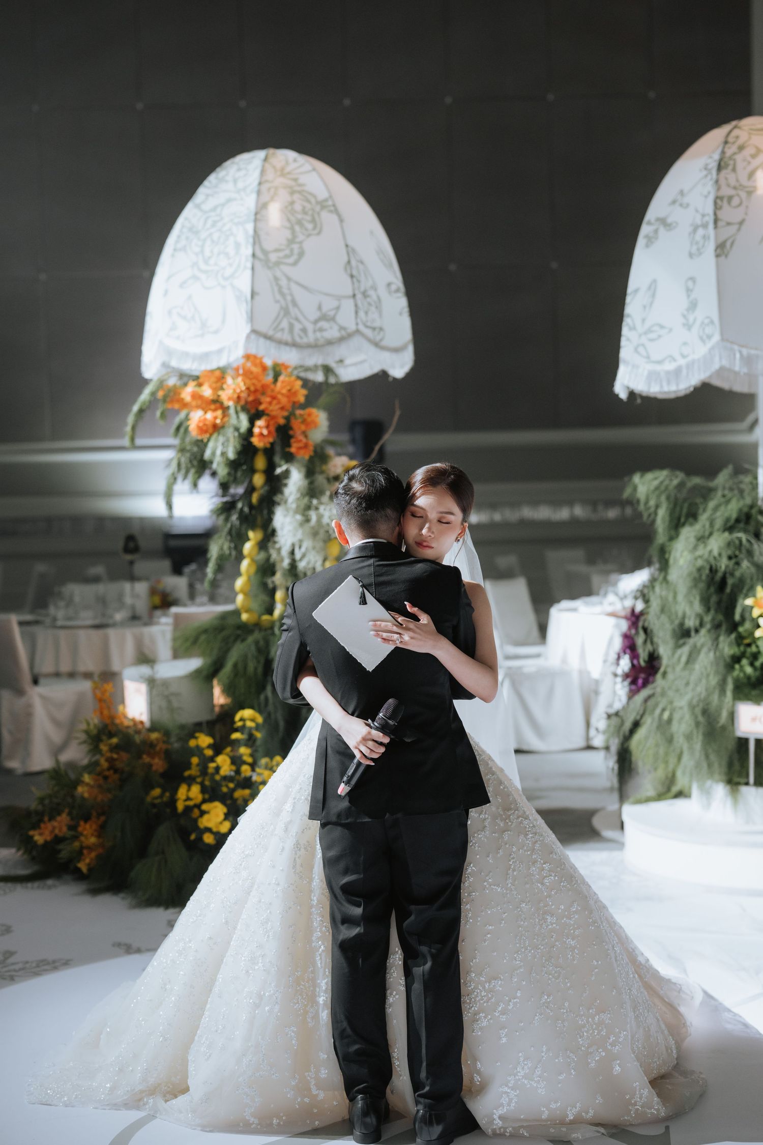 A couple embraces at their wedding ceremony under elegant white lace lampshades surrounded by orange and yellow florals.
