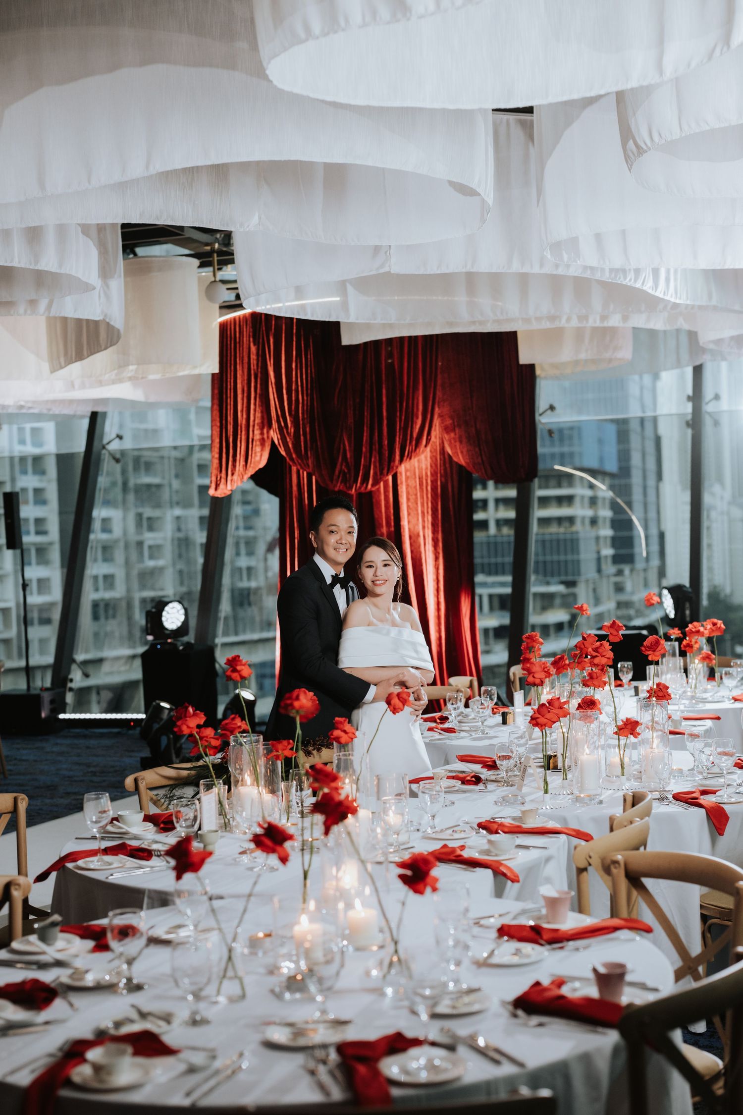 Modern wedding reception table with elegant white linens, red floral centerpieces and romantic candlelight under white draped ceiling.