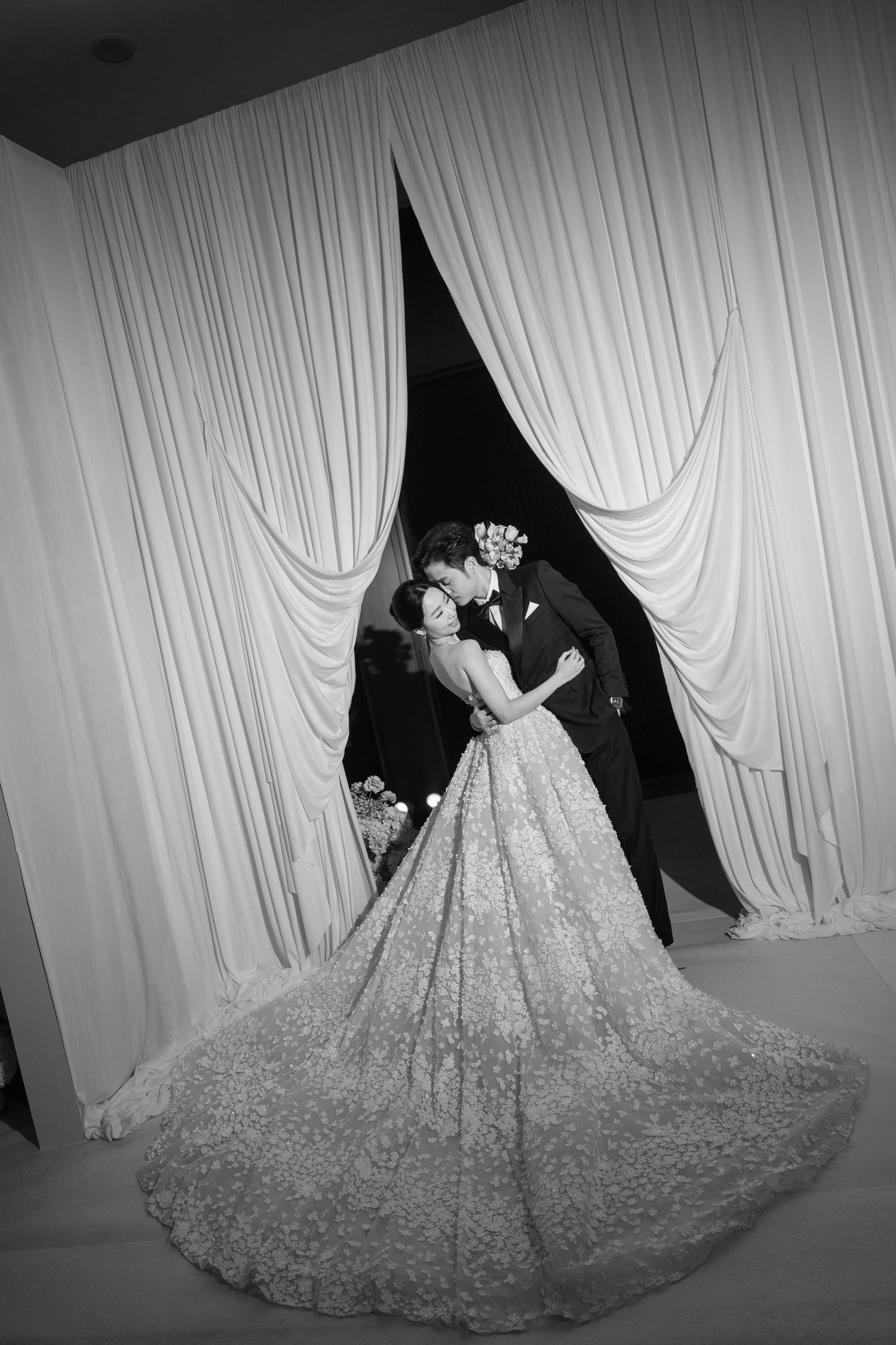 Black and white wedding photo of bride and groom kissing between elegant draped curtains, bride in stunning lace ballgown.