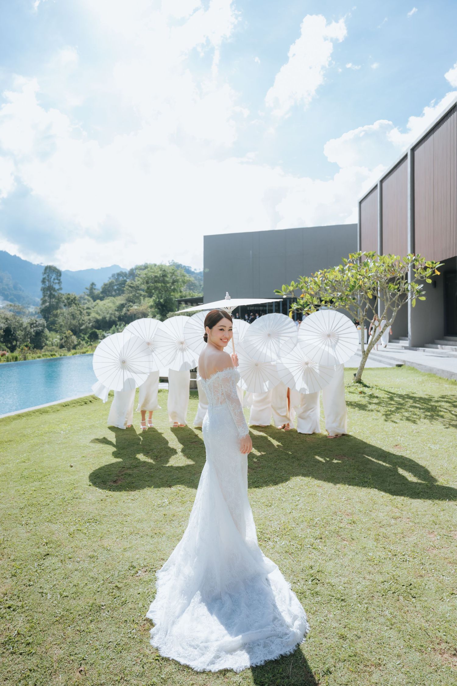 Elegant bride in white lace gown poses outdoors on green lawn with white parasols and mountain landscape in background.