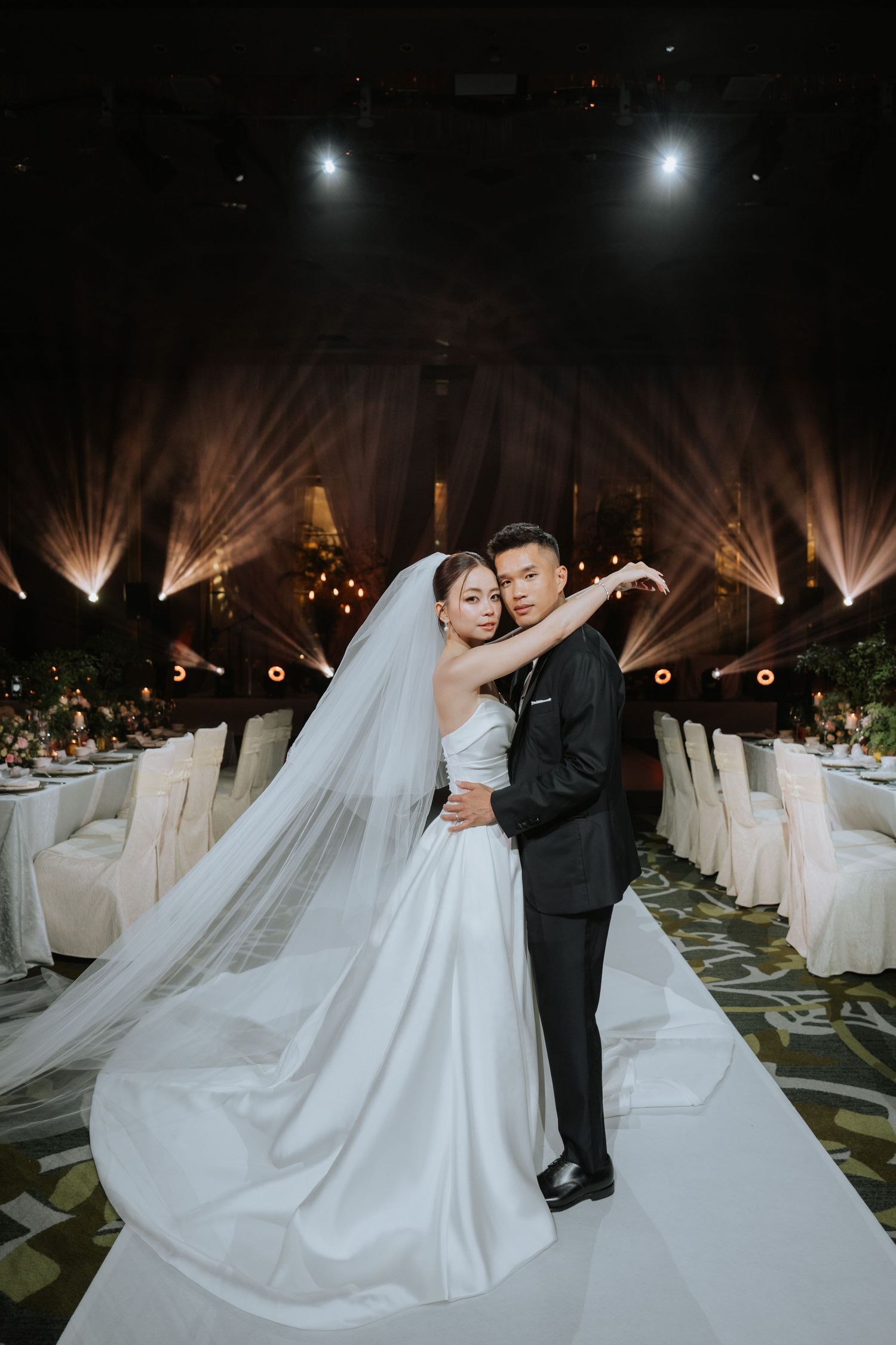 A wedding couple embraces on a white aisle runner in an elegant ballroom with dramatic uplighting and lined chairs.