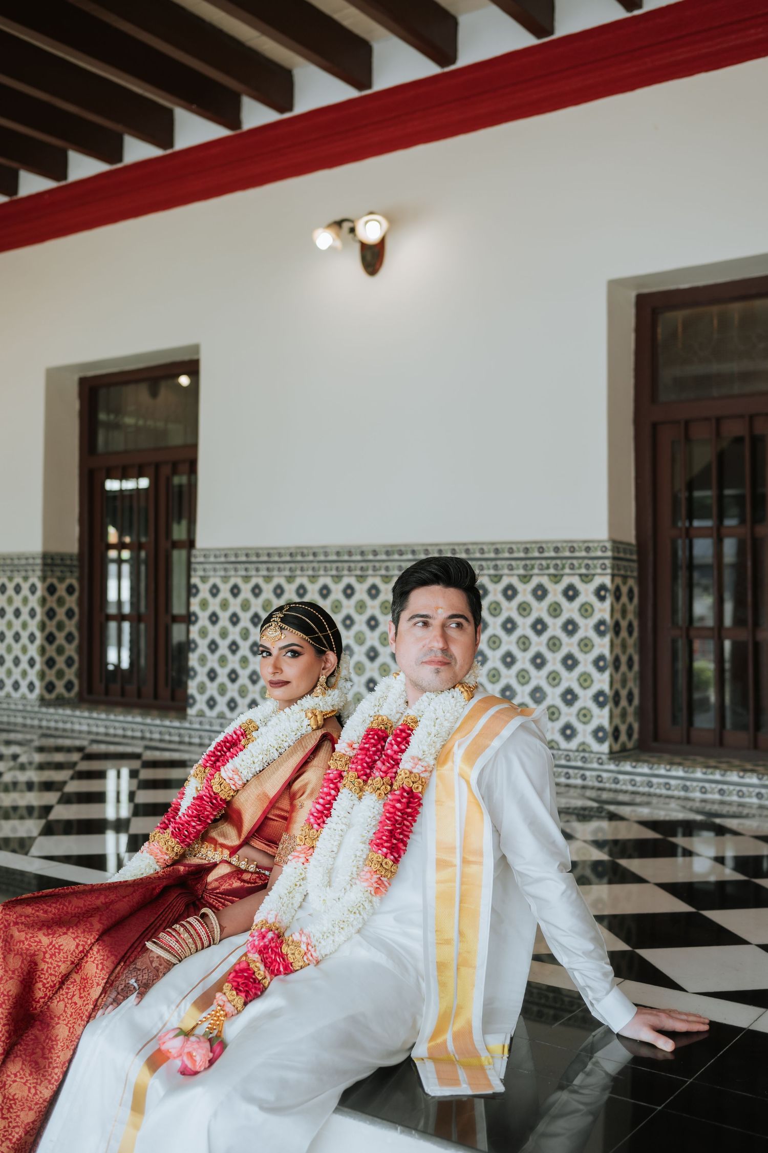 Indian bride in red saree and groom in white kurta wearing floral garlands seated together at a traditional wedding ceremony.